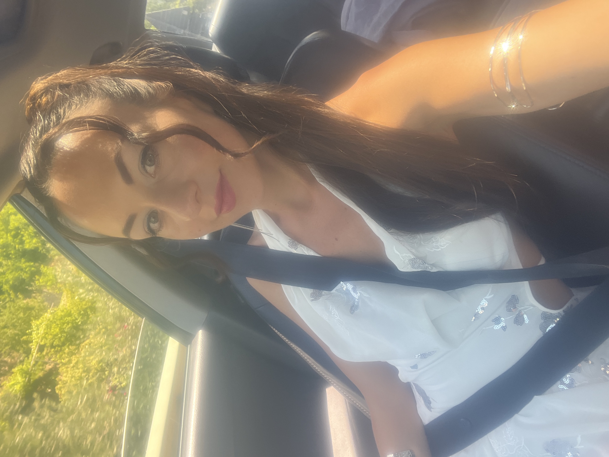 A woman is sitting in the passenger seat of a car, with her seatbelt fastened across her chest. She is wearing a white dress with a sequined pattern and ornate silver bracelets on her upper arms. Her hair is pulled back into a ponytail, with loose strands framing her face. The car appears to be moving, as indicated by the blurred background of trees and foliage, suggesting a scenic route, possibly near Đurmani, Montenegro. The sun is casting a warm glow on the scene, highlighting the woman's features and the details of her jewelry and dress.