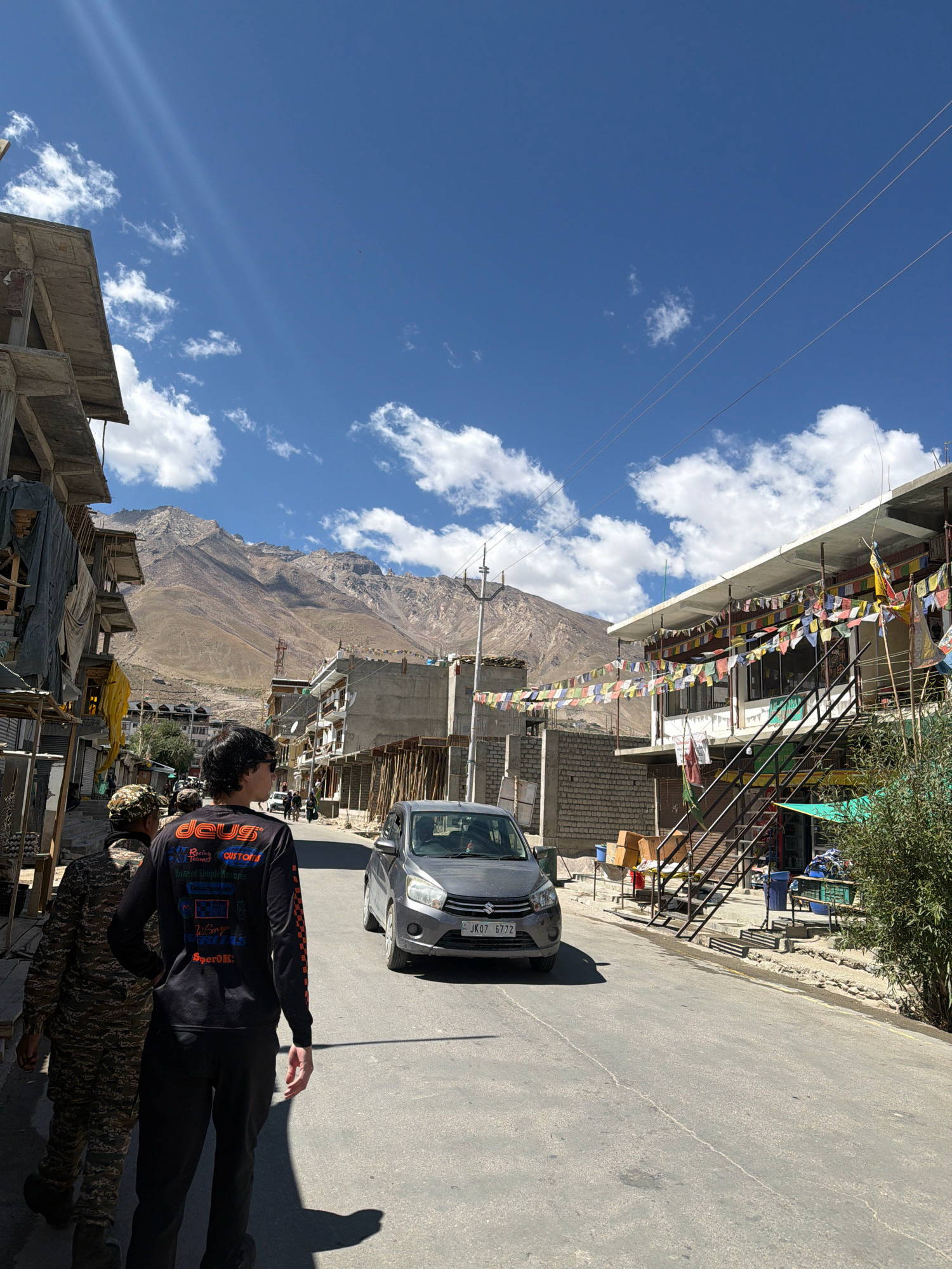 In Padum, India, a street scene unfolds under a bright blue sky with scattered white clouds. A man wearing a black long-sleeved shirt with colorful graphics and black pants walks away from the camera, casting a shadow on the asphalt road. Beside him, a person in camouflage fatigues walks in the same direction. A grey car with license plate "JK07 6772" is visible on the road. Buildings line both sides of the street, some with balconies adorned with colorful prayer flags. In the background, a large, rocky mountain looms under the clear sky. The architecture suggests a town in a mountainous region, likely in a region like Ladakh or Himachal Pradesh, given the prayer flags and the dry, mountainous landscape, consistent with Padum in the Zanskar valley.