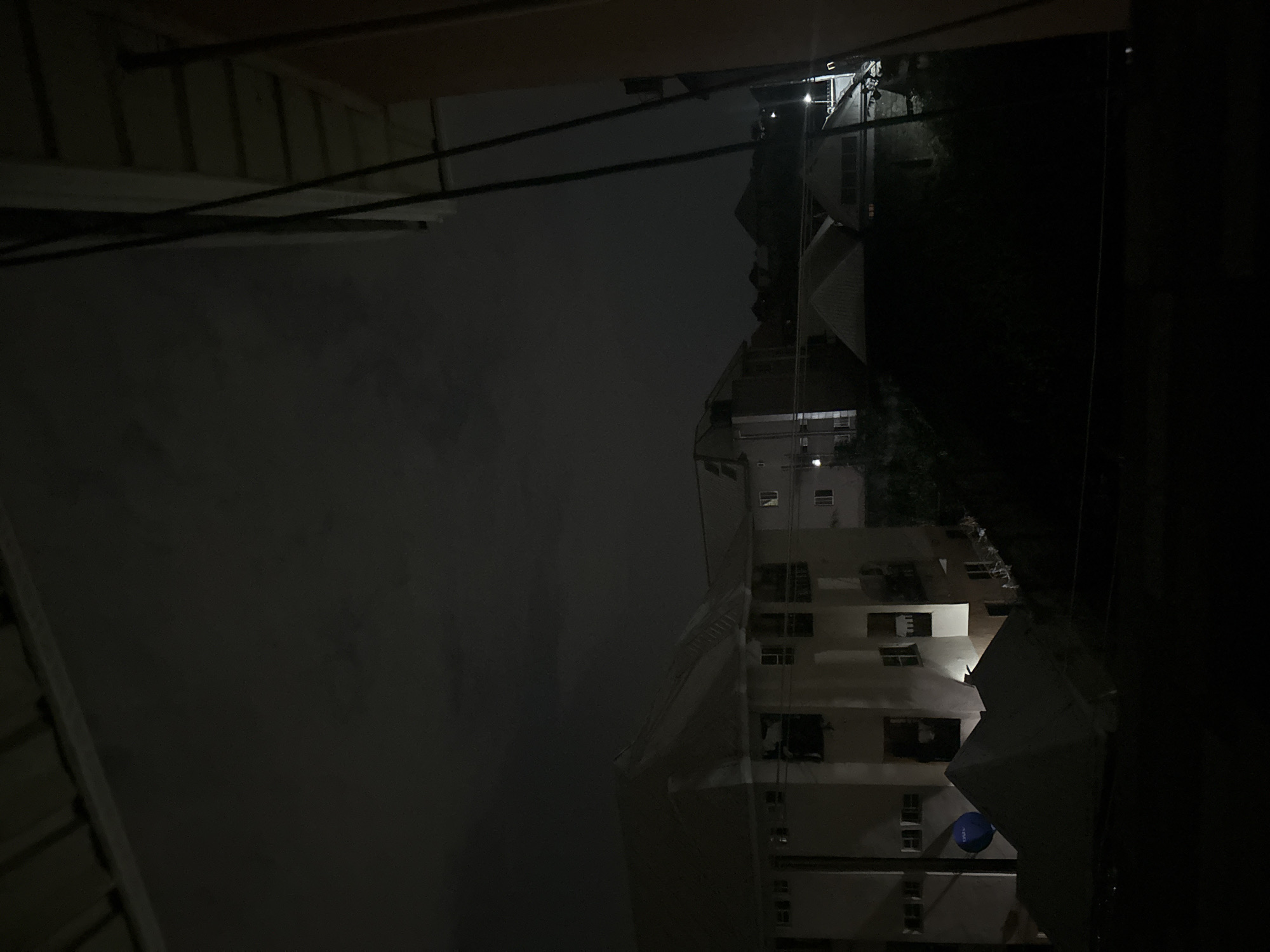 At night in Ihiagwa, Nigeria, a tall, light-colored building stands illuminated. Scaffolding partially obscures the facade. To the left, a darker building is visible. The sky is overcast. Electrical wires run across the scene.