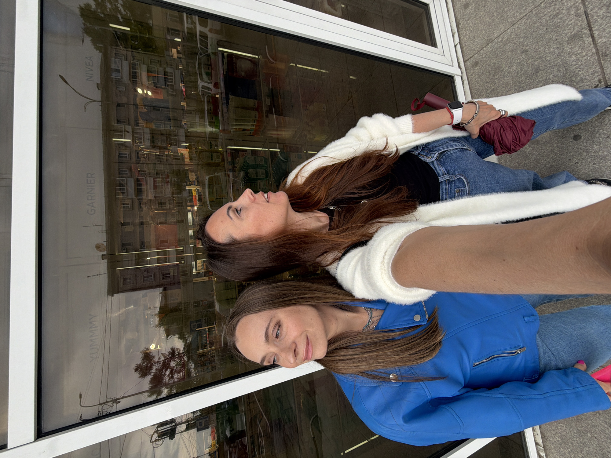 Two women are posing in front of a storefront window. The woman at the top is wearing a white cardigan over a black top and blue jeans. The other woman is wearing a blue leather jacket and blue jeans. The storefront window reflects buildings and street elements. The storefront has brands "GARNIER" and "NIVEA" visible on its signage. Another label, "YUMMY," is also visible in the reflection. The women are likely in a city; while it is difficult to confirm the location, the reference to Birobidzhan, Russia, suggests it could be there or a similar location.