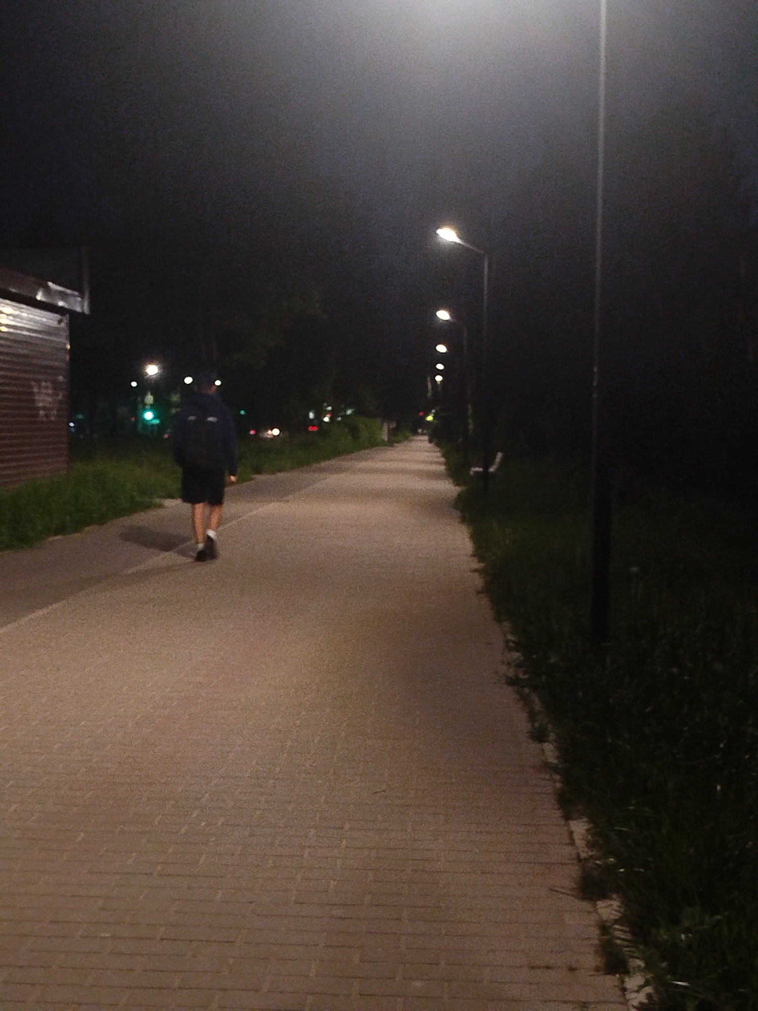 The image shows a paved walkway at night, illuminated by a series of streetlights. A person wearing a dark hoodie, dark shorts, and white socks with dark shoes, is walking away from the viewer, down the walkway. The walkway is flanked by grassy areas on either side. A small building with corrugated metal walls is on the left. In the distance, there are faint streetlights and possible traffic lights. The location is in an urban park.
