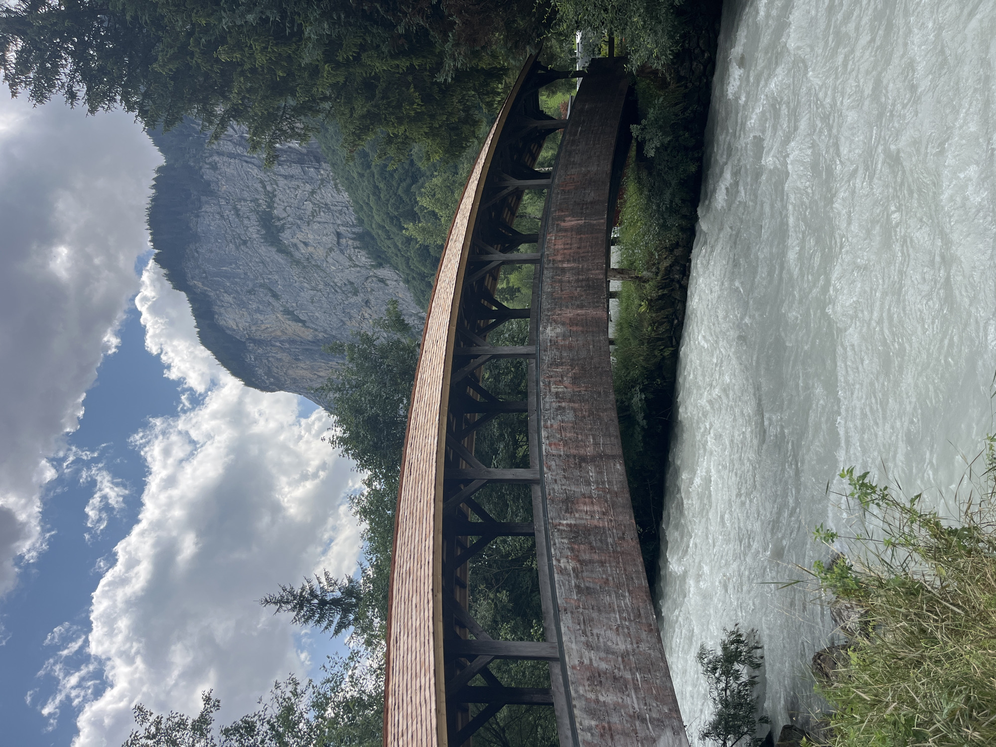 This is a full, eye-level, outdoor, vertical shot of a covered wooden bridge arching over a fast-flowing river. The bridge has a reddish-brown wooden roof and sides, with exposed wooden support beams visible underneath. The river is a milky, turbulent white, indicative of glacial meltwater, and runs from the bottom left to the middle right of the frame. The riverbanks are lined with green grass and foliage. In the background, a large, sheer, gray rock face rises dramatically, topped with a dense evergreen forest. The sky above is blue with scattered white, puffy clouds, and dappled sunlight. The scene is likely located in Wengen, Switzerland, given the characteristic mountain landscape and architecture. There are no people visible in the image.