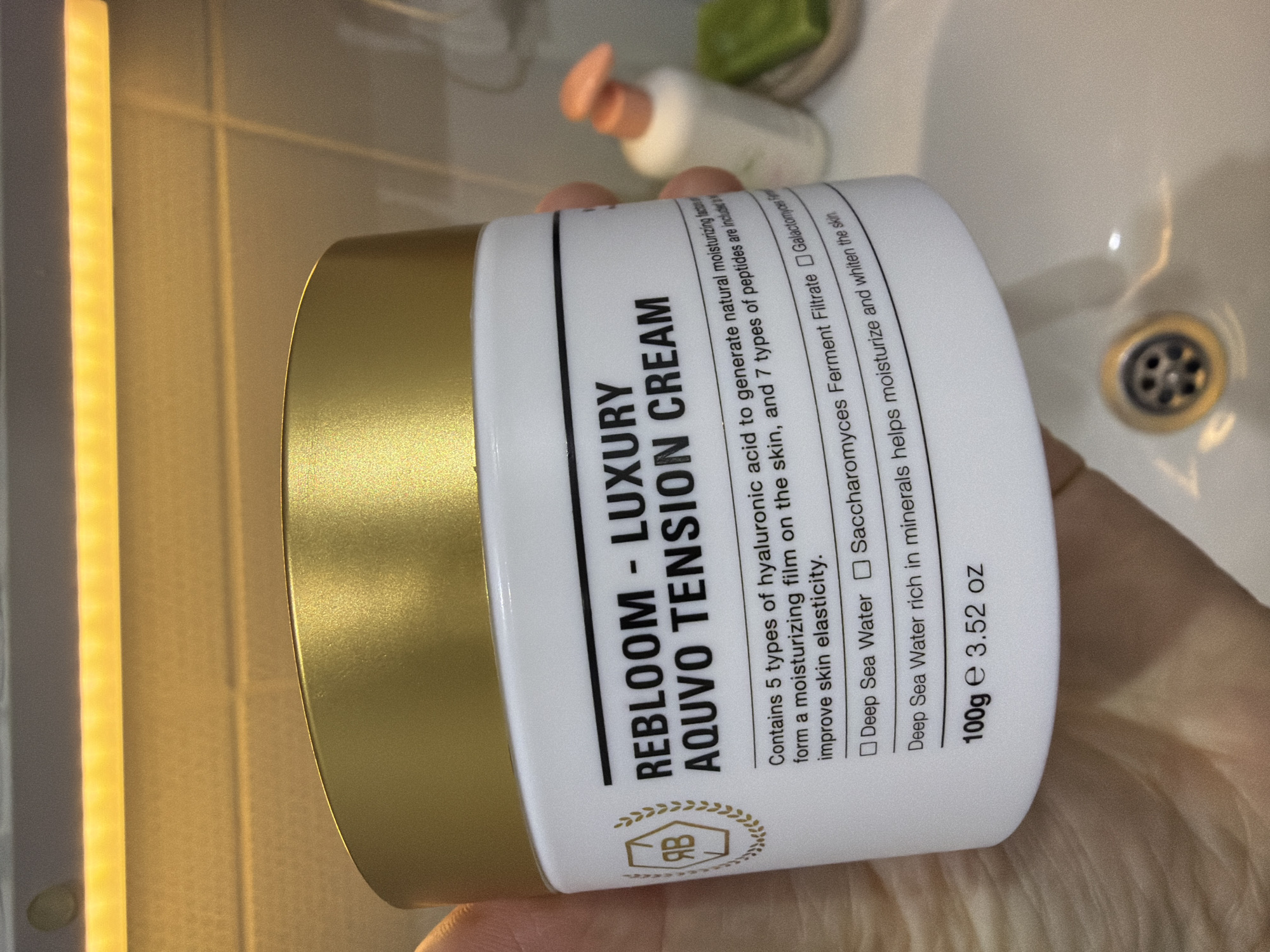 The image shows a white jar with a gold lid, labeled "REBLOOM - LUXURY AQUVO TENSION CREAM". The text on the label indicates the cream contains hyaluronic acid, peptides, deep sea water, and Saccharomyces Ferment Filtrate. The jar is held in someone's hand, and in the background is a bathroom sink with a drain, a partially visible bottle, and a tiled wall with a light fixture. The location in Chaikovsky, Russia is not discernible from the image.