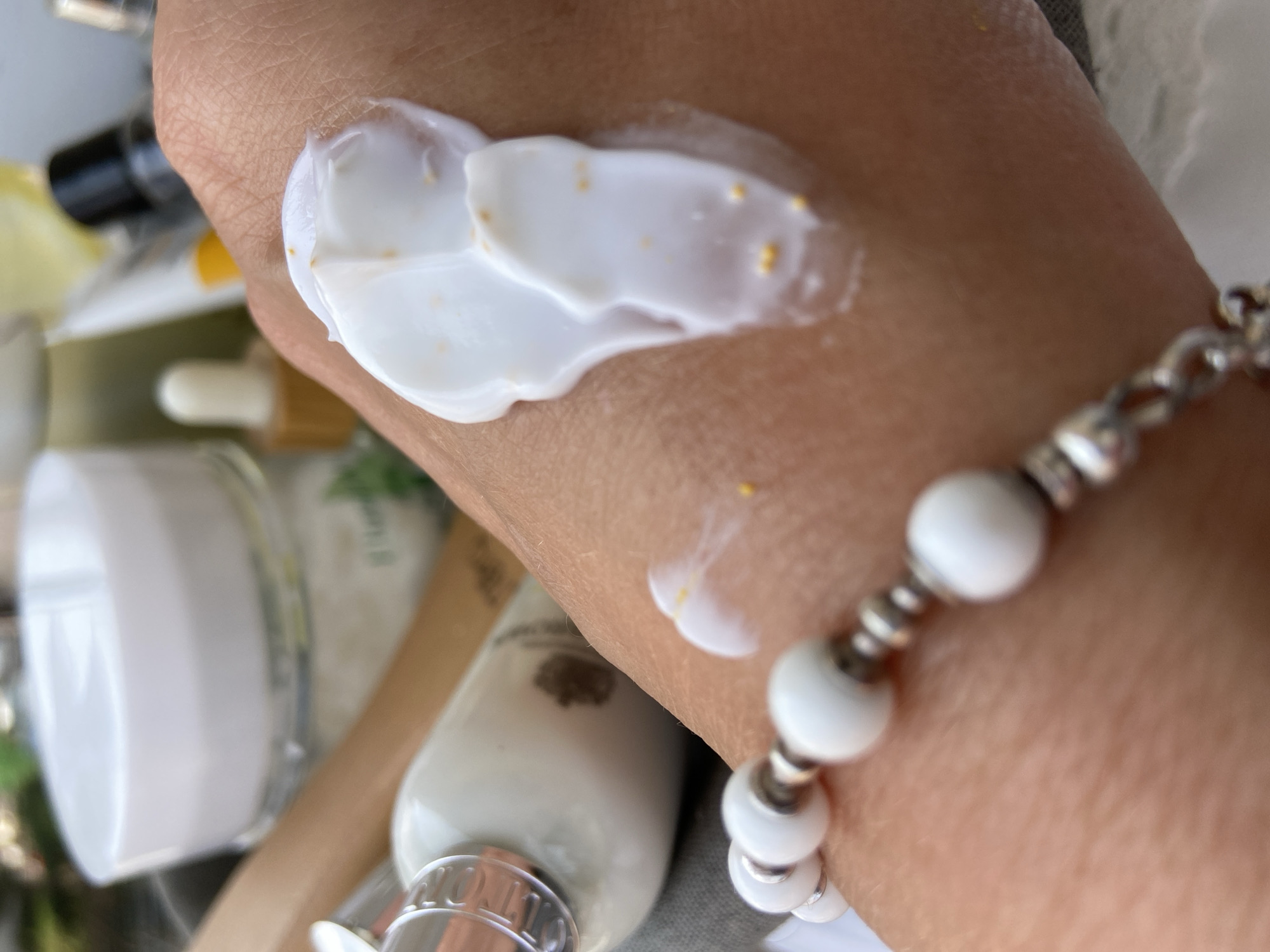 A close-up shot depicts a caucasian hand with white cream and gold specks applied to the skin. A white beaded bracelet adorns the wrist. Several beauty product containers of various shapes and sizes are blurred in the background. A small amount of what appears to be the same white cream is on the forearm closer to the wrist. The location and time of day cannot be determined from the image.