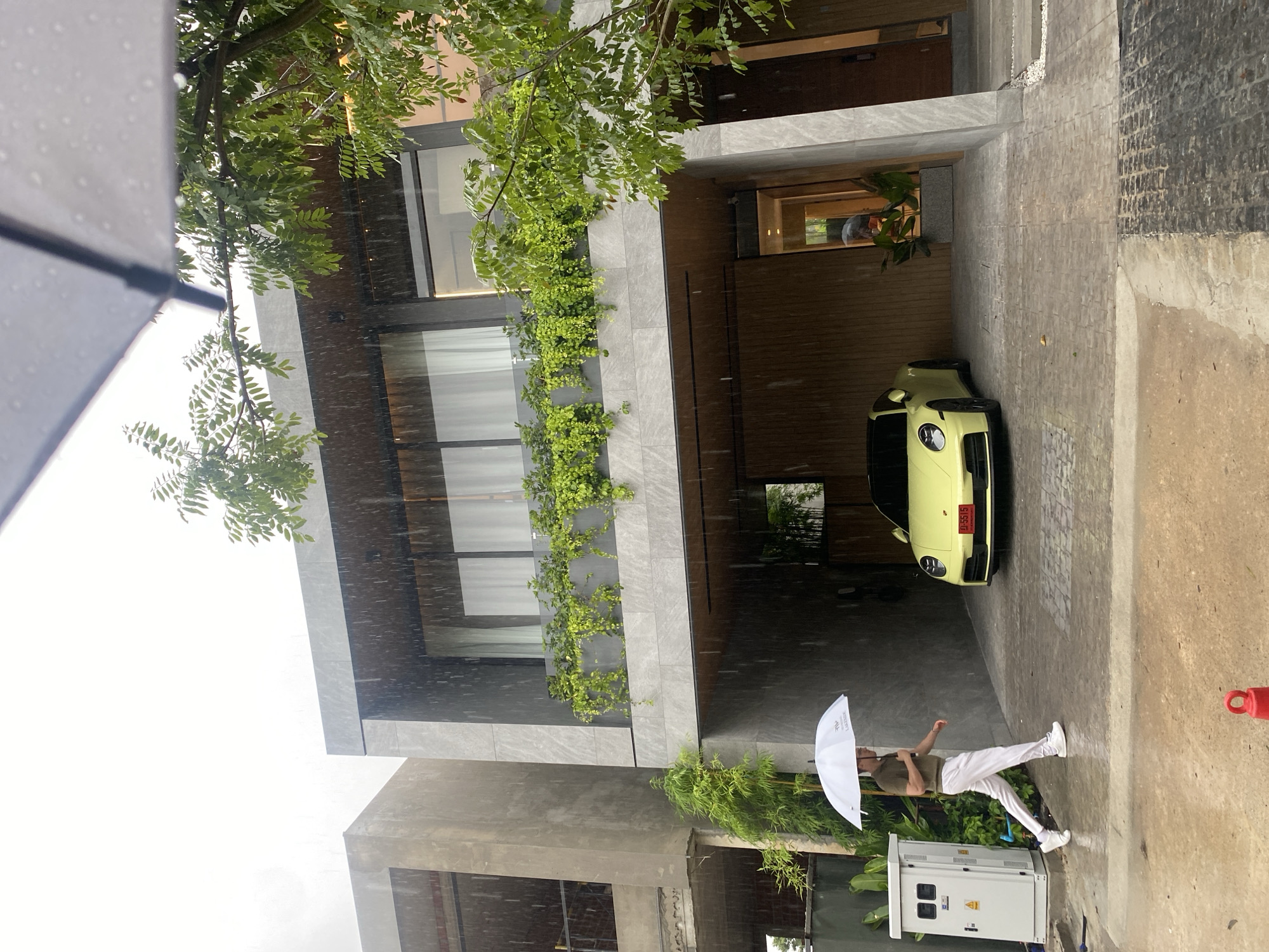 On a rainy day in Choeng Thale, Thailand, a modern two-story building stands with greenery draping its façade. A light yellow sports car is parked in the building's carport. A person holding a white umbrella walks on the sidewalk near an electrical box. The scene conveys a sense of urban luxury combined with tropical foliage. Rain is visible throughout the shot.