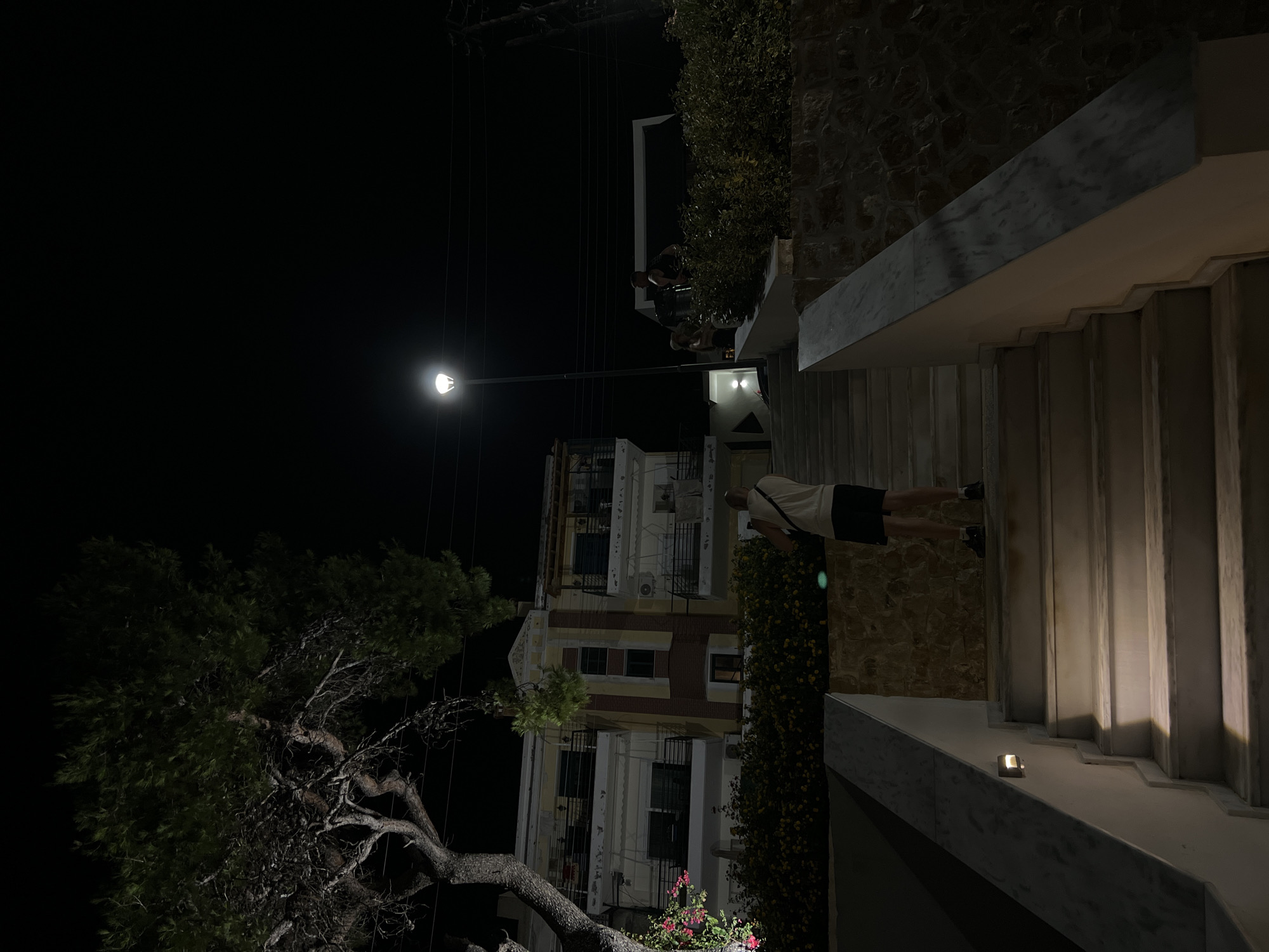The image depicts a nighttime scene with a person ascending a marble staircase. The staircase is illuminated by built-in lights and a distant streetlight. To the left of the staircase, there is a building with multiple balconies, and dense foliage with some flowering plants. The background is dominated by the night sky and a street lamp. The environment appears to be a public outdoor space, possibly a residential area or a commercial establishment within Sithonia Municipal Unit, Greece. The person is wearing a white tank top and black shorts, carrying a bag across their chest. Other individuals are visible further up the stairs and on a balcony of the building. The overall atmosphere is tranquil and illuminated.