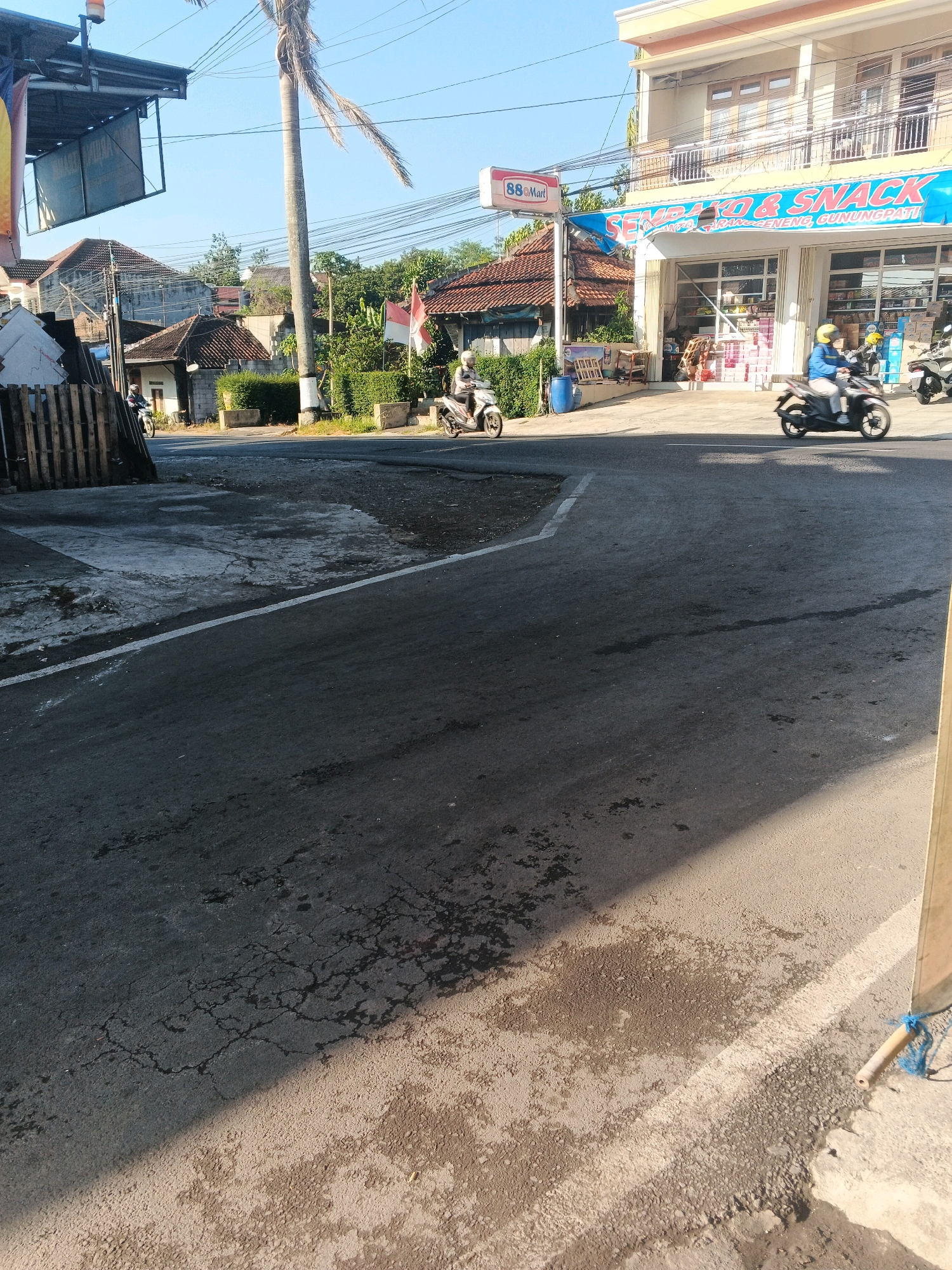 The image captures a street scene in the city of Semarang, Indonesia, likely in a residential or commercial area. The foreground is dominated by a dark asphalt road with some patches of wetness and cracks, indicating recent rain or a water spill. A white line marks the edge of the road. On the right side of the frame, a portion of a light-colored building or structure is visible, with a blue rope tied to a bamboo pole.
In the mid-ground, a multi-story building with a beige facade and balconies stands prominently. A blue banner with white text, reading "SEMBAKO & SNACK" and other Indonesian words, is displayed across the front of the building, suggesting it is a convenience store or a shop selling groceries and snacks. A sign for "88 Mart" is also visible on a pole next to the store.
Several people on motorcycles are present on the street. Two individuals are riding motorcycles from left to right. One person is wearing a white helmet and appears to be on a white scooter. Another person, further to the right and closer to the store, is wearing a yellow helmet and a blue jacket, riding a black scooter. A third motorcycle is partially visible in the background to the far right.
The background shows a cluster of houses with red tile roofs and some greenery, including a tall palm tree with sparse fronds. Indonesian flags are visible on some of the houses. The sky is clear and blue, suggesting a sunny day. The overall impression is of a typical street in a lively Indonesian neighborhood.