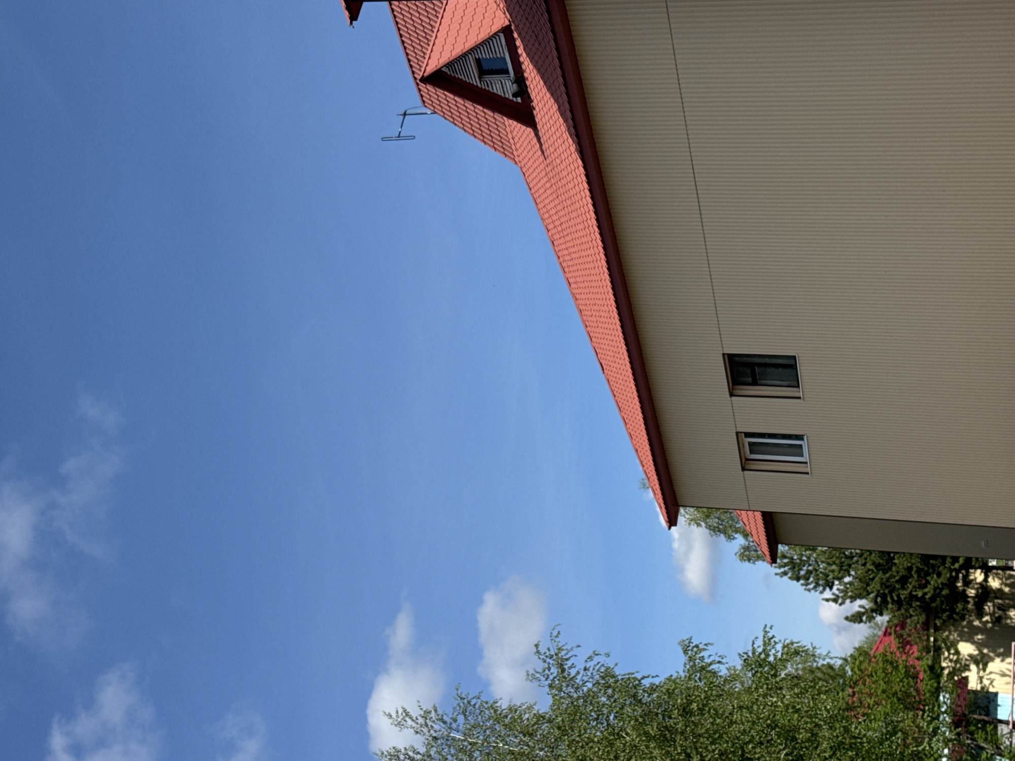 The image displays an outdoor scene, likely in Высокий, Russia, under a clear, bright blue sky with scattered white, fluffy clouds. The primary subject is a building, viewed from a low angle, which features a vibrant red tiled roof with a textured pattern. A striking triangular dormer window is integrated into the roof, visible on the upper left side of the main roof slope. Adjacent to this, a metallic, antenna-like structure extends horizontally from the roof.
The building's facade is covered with light beige or tan corrugated siding, and two rectangular windows are visible on this side. A thin wire or cable runs diagonally across the siding in one section. Lush green trees with dense foliage occupy the lower and bottom-left portions of the frame, partially obscuring another building with a red roof in the bottom-right corner. The scene appears to be set during daytime, with ample sunlight. There are no people or specific activities taking place, and no discernible text is visible within the image. The overall impression is that of a quiet, clear day in a residential or built-up area.