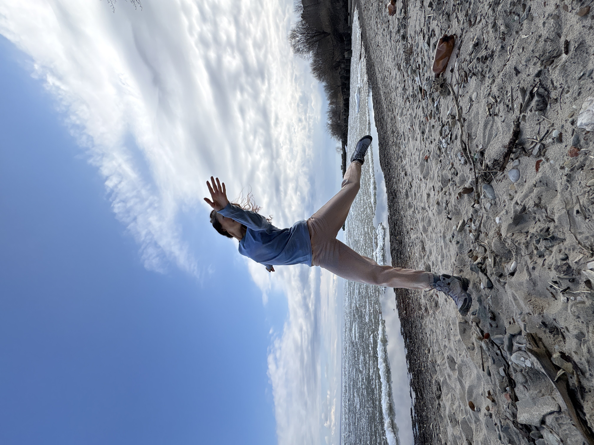 A woman is performing a yoga pose on a sandy beach next to a body of water. The sky is partly cloudy with a gradient from blue to white. The beach surface consists of sand mixed with pebbles and driftwood. A tree line is visible in the background. The image is rotated, making it appear as if the woman is suspended in mid-air. There is no confirmed information about the city of Ulyanovsk, Russia, in the image to confirm this location.