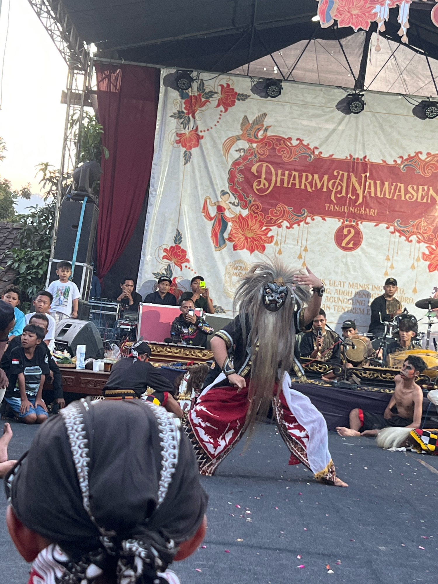 In Madyocondro, Indonesia, a performance is underway on an outdoor stage. A performer wearing a mask and elaborate costume with a long, wild wig is dancing in the foreground. The mask is grey and black with red accents and large, protruding teeth. The costume includes a black top with gold trim and a flowing red and white skirt. Behind the dancer, a band is playing traditional Indonesian instruments, including drums and what appears to be a gamelan. Several individuals are seated behind the band, watching the performance. In the background, a large banner with red and gold floral designs and Indonesian text reads "DHARMNAWASEN TANJUNGSARI" and a circled number "2." The stage is lit by spotlights, and a large speaker is visible to the left. A crowd of spectators, including children, are visible in the foreground, some looking towards the stage and others interacting amongst themselves. The ground is paved and scattered with fallen flower petals.