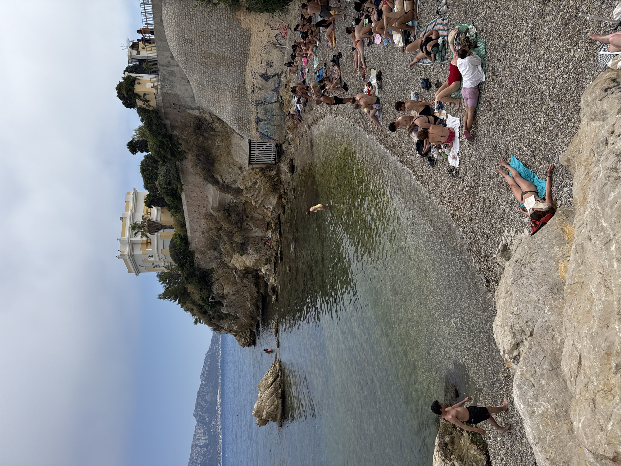 The image shows a rocky beach in Marseille, France, with a cliffside building and the Mediterranean Sea in the background. A man in black swim trunks is walking along the pebble beach towards the water. Several people are sunbathing on towels scattered on the beach. Some are lying down, while others are sitting. The water is clear and shallow near the shore, with a large rock formation in the sea. In the distance, mountains and a city skyline are visible. The sky is overcast.
