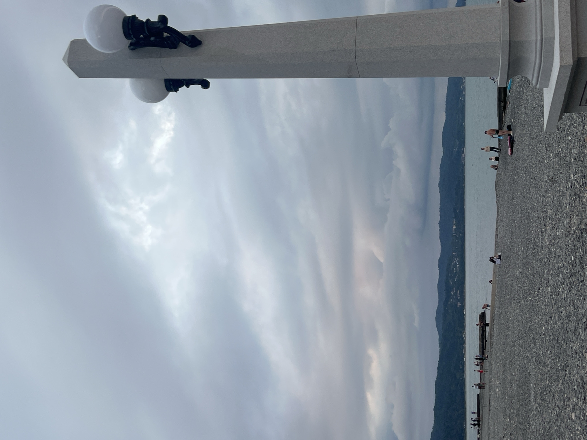 This image depicts a cloudy day at a pebble beach in Sokhumi, Abkhazia. In the foreground, a tall, grey stone lamppost with two globes stands to the right. The beach is covered in grey pebbles, and a few people are scattered across it and near the water's edge. Some individuals are standing on long, dark structures that extend into the sea, possibly piers or breakwaters. The sea is calm, and in the background, lush green hills rise, dotted with buildings, forming the coastline of Sokhumi. The sky is overcast with heavy grey clouds.