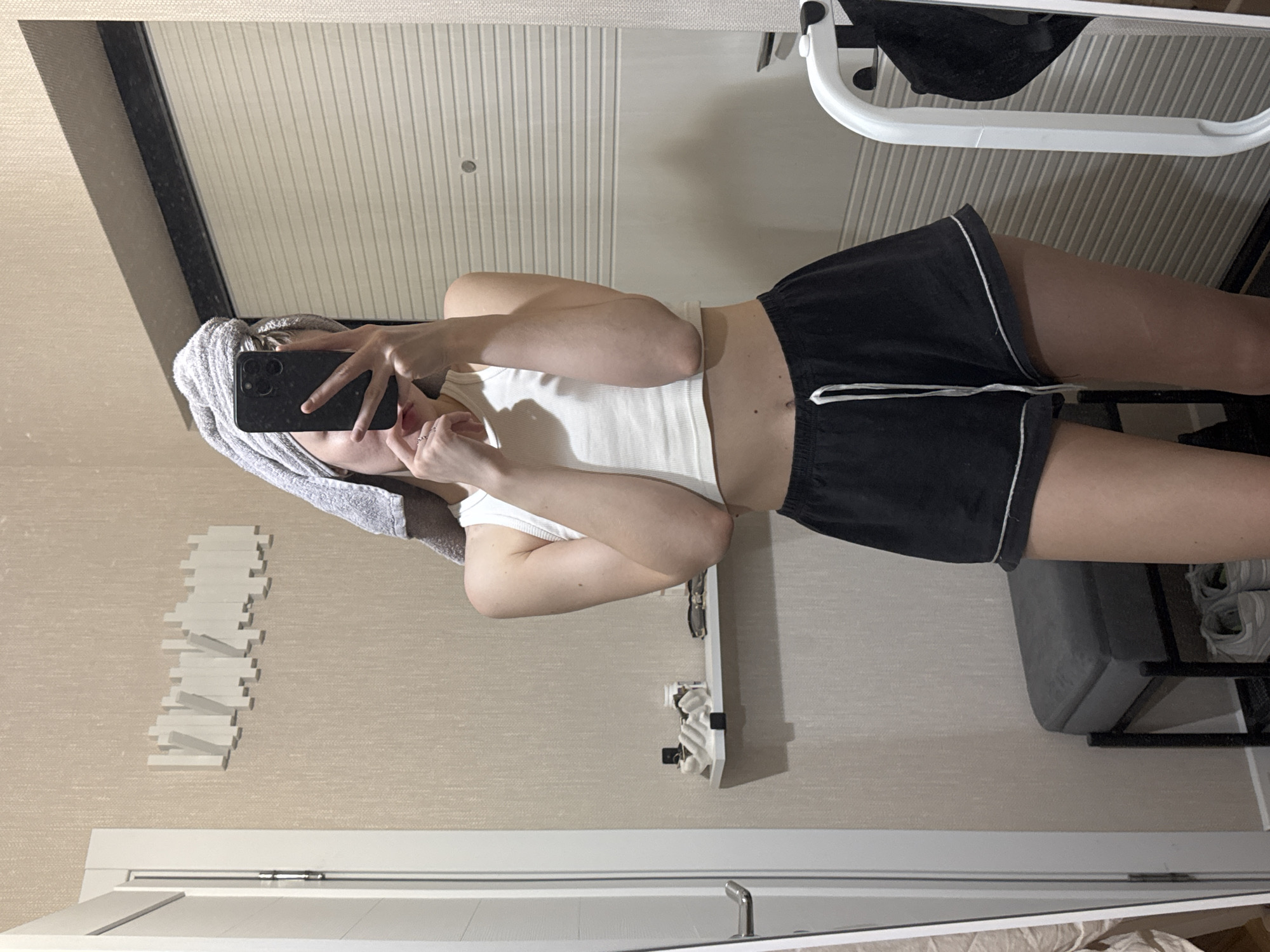 A woman is taking a selfie in a room. She has a towel on her head, is wearing a white tank top and black shorts. There is a white rack on the wall with a white storage rack below. A folding chair is visible in the background. The location is not apparent from the image.