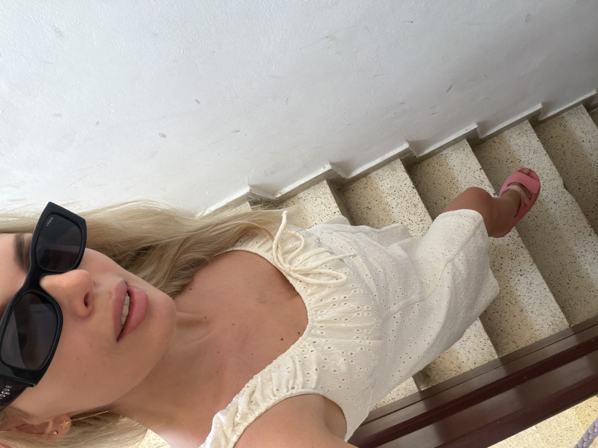 A young woman with blonde hair and light skin is standing on a staircase. She is wearing black sunglasses and a white eyelet dress. She has pink lipstick on and her mouth is slightly open. Her right foot is visible, wearing a pink sandal. The staircase has speckled stone steps and a brown wooden railing. The wall next to the staircase is white. The scene appears to be outdoors, possibly in a sunny location, given the lighting. The overall mood seems casual and relaxed. Visible text on the sunglasses reads "VOGUE".