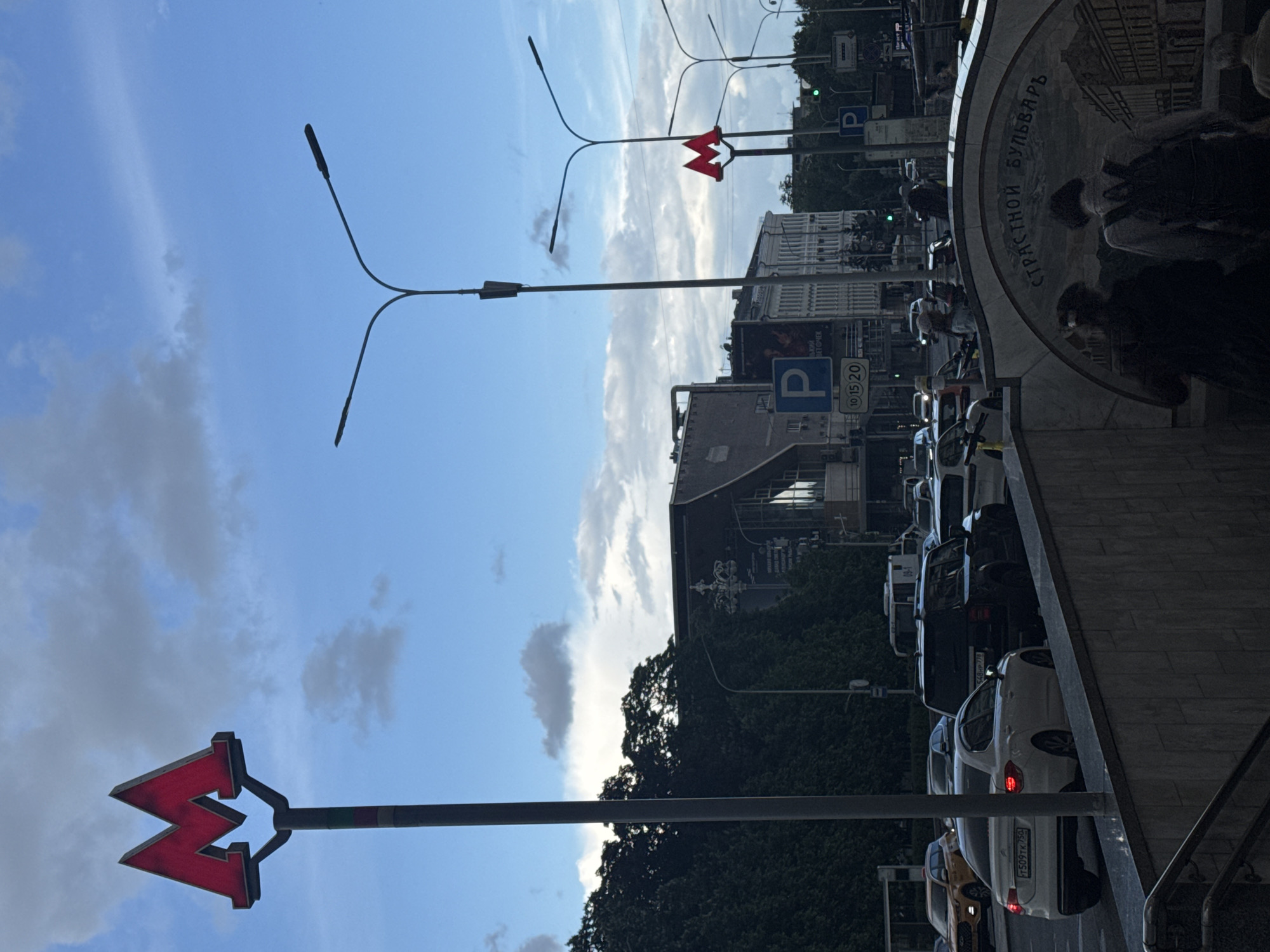 The image depicts a busy urban street scene in Moscow, Russia, likely during the late afternoon or early evening under a partly cloudy sky. The perspective is from a slightly elevated position, looking down a street filled with traffic.
The primary subjects are numerous cars, appearing to be in a traffic jam or moving slowly. Buildings of various heights line both sides of the street. Prominently featured are two red "M" signs, emblematic of the Moscow Metro, indicating nearby subway stations. One sign is on a tall pole in the foreground on the left, and another is further down the street on the right.
Activity centers around the flow of urban traffic and pedestrian movement near the Metro entrance. On the right side of the image, a distinctive semicircular decorative panel, part of a Metro entrance, is visible. This panel features the Cyrillic text "СТРАСТНОЙ БУЛЬВАРЪ" (Strastnoy Boulevard), confirming the specific location. Several people are partially visible near this entrance.
Notable details include streetlights with multiple arms, parking signs displaying a blue "P" symbol along with numbers "10 15 20," and Russian license plates on some vehicles, such as "T509TK 750" and "548PB 777." Trees and foliage are interspersed among the buildings, and a clear blue sky with scattered clouds forms the backdrop. The scene captures a typical moment of urban life and commuting in the Russian capital.