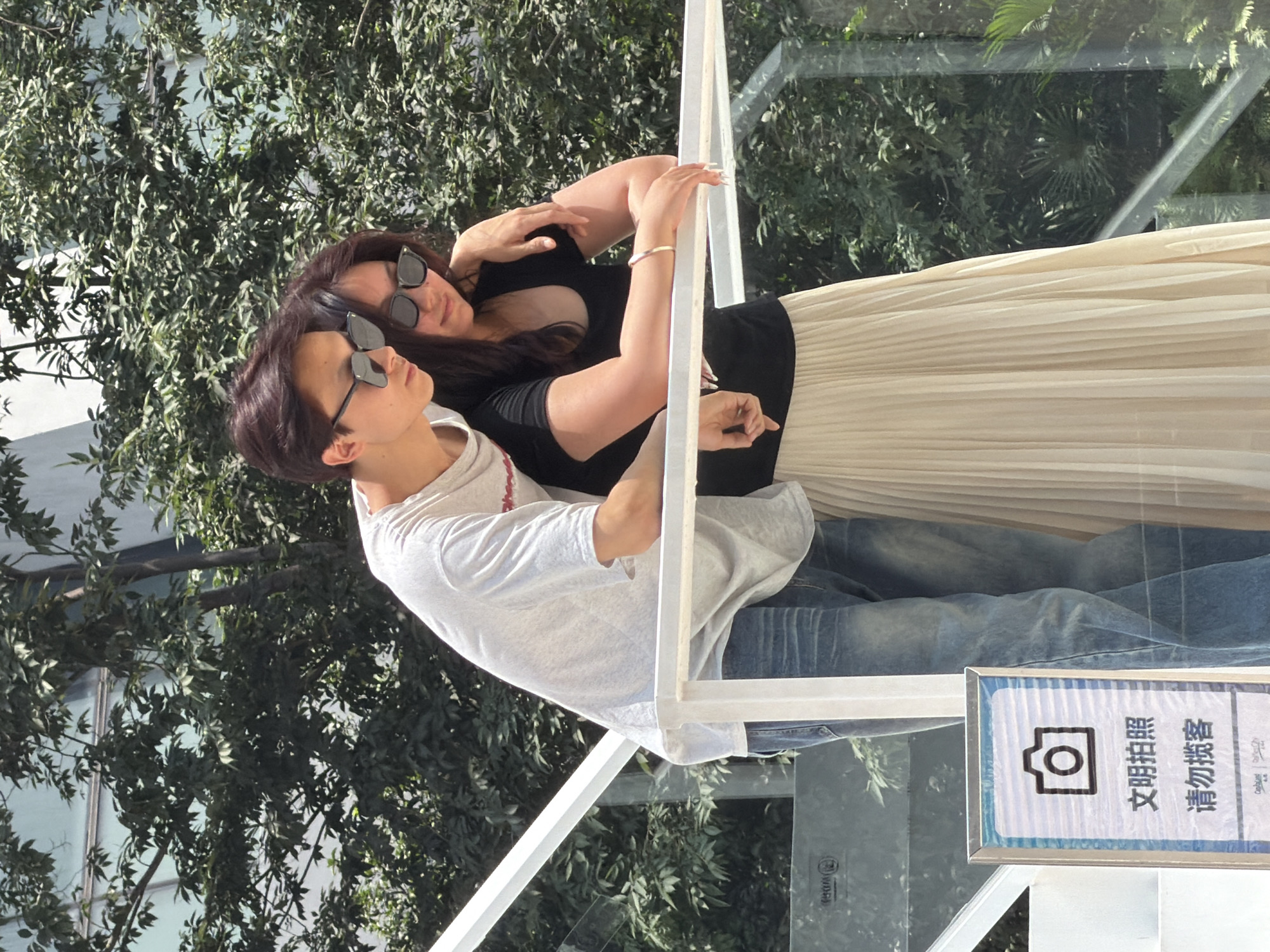 In Yuzhong District, China, a young man and woman are standing outdoors near a white railing. The man, wearing a light-colored t-shirt and blue jeans, has his arm around the woman's shoulders. The woman, dressed in a black t-shirt and a long, cream-colored pleated skirt, leans into him, her arm resting on the railing. Both are wearing sunglasses. Behind them, lush green trees and foliage are visible, with a modern building in the background. To the left of the couple, a sign with a camera icon and Chinese text is mounted on a white stand. The overall setting appears to be an elevated walkway or balcony.