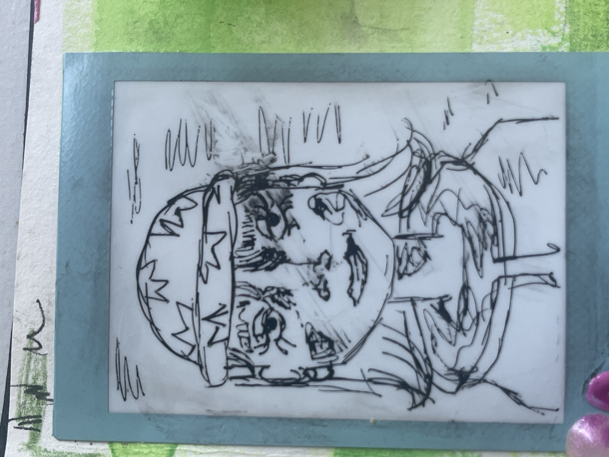 The image displays a framed, black-and-white line drawing, presented horizontally but depicting figures that appear upright when rotated 90 degrees clockwise. The artwork is set on a mixed media background of textured white paper and a green painted surface, with additional decorative elements at the bottom right.
The central subject is a stylized, expressive drawing featuring two human-like faces or busts in close proximity. The figure on the left side (when upright) has a head covering adorned with zigzag patterns, large wide eyes, and a simple mouth. The figure on the right side also features large eyes, a distinct nose, and an open mouth revealing teeth, conveying an animated expression, possibly of surprise or excitement. The drawing is executed with bold, fluid black lines and exhibits some smudges, suggesting a spontaneous sketch or ink transfer.
The drawing is contained within a light blue, rectangular frame, which appears to be made of cardstock with a slight texture. This framed piece rests on a background that includes a section of white, textured paper or canvas on the left, and a vibrant green, painted or textured surface covering the top and right portions. In the bottom right corner of the overall image, outside the blue frame, are several small, shiny, irregularly shaped pinkish-purple objects, resembling beads or decorative stones.
There is no legible text visible within the drawing or on the frame; only abstract, vertical scribbles appear within the drawing's upper section. The overall scene is brightly lit, emphasizing the textures of the various materials.