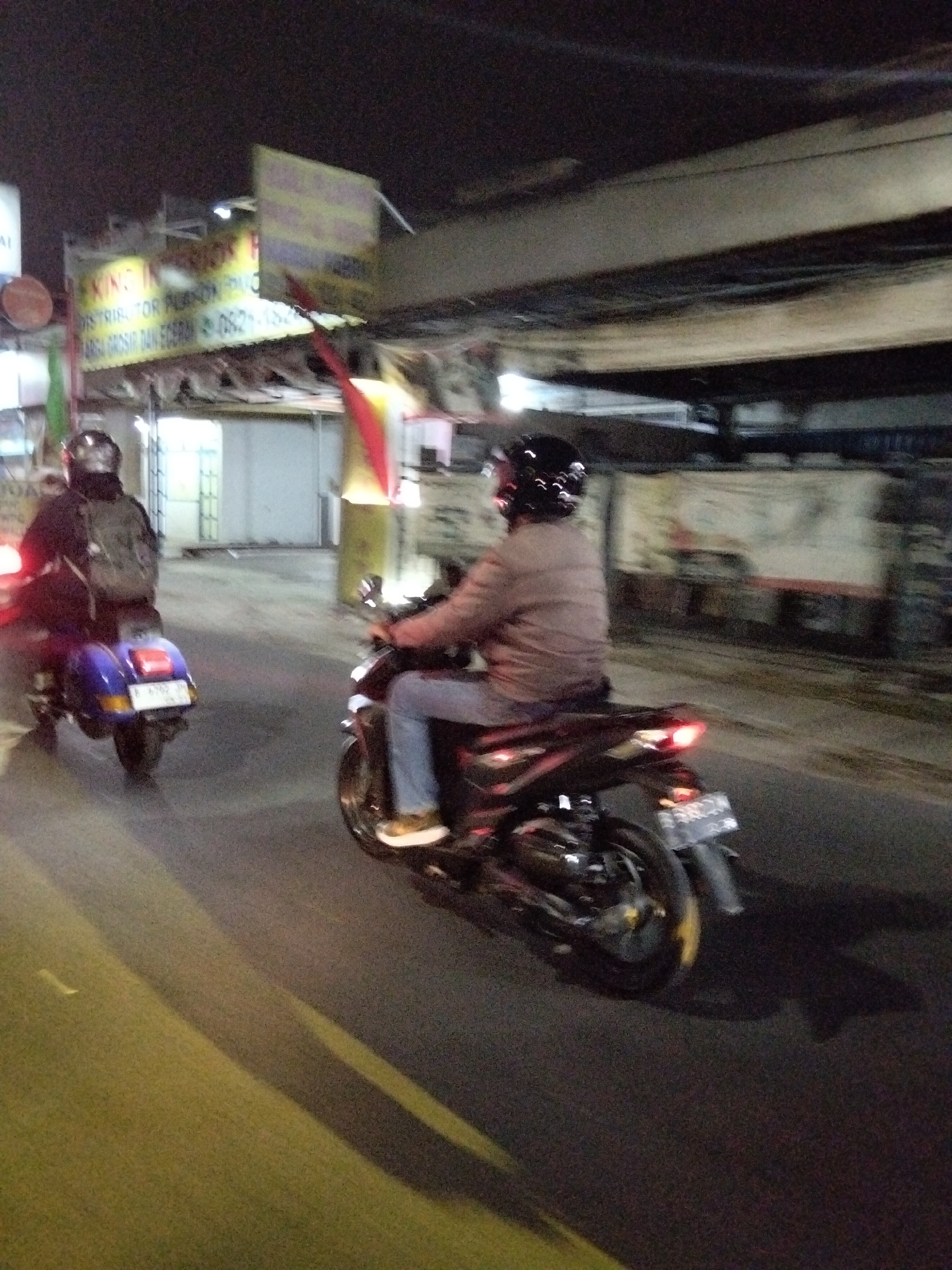 An outdoor, night-time, slightly blurry, medium shot depicts two people riding motorbikes on a paved road in South Tangerang, Indonesia. The person in the foreground is on a dark-colored motorbike, wearing a light brown jacket and blue jeans, and a black helmet. The person in the background is on a blue motorbike, wearing a dark jacket and a backpack, and a dark helmet. Yellow lines are visible on the road, indicating lanes or a crosswalk. A street light casts a warm glow on the scene. In the background, there are buildings and a sign with Indonesian text that is partially obscured.
