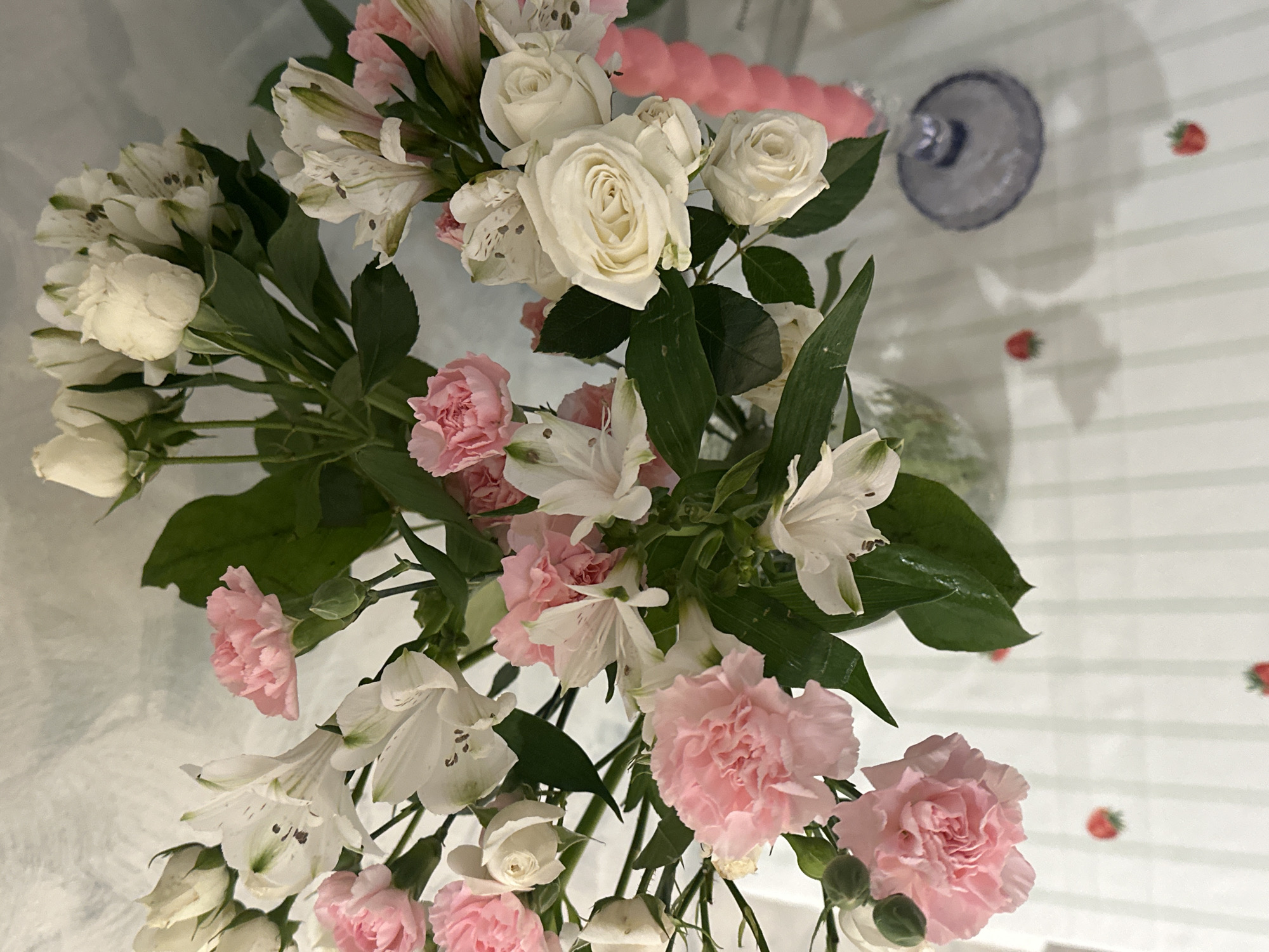 A high-angle, close-up shot captures a bouquet of flowers against a white surface with a light gray, striped pattern, and strawberry decorations. The flowers include white roses, pink carnations, and white alstroemeria with green foliage. A partially visible clear glass object rests in the upper right corner. In the upper background, there is a pink object. The scene appears indoors, lit by ambient lighting. The location is not discernible from the visual information.