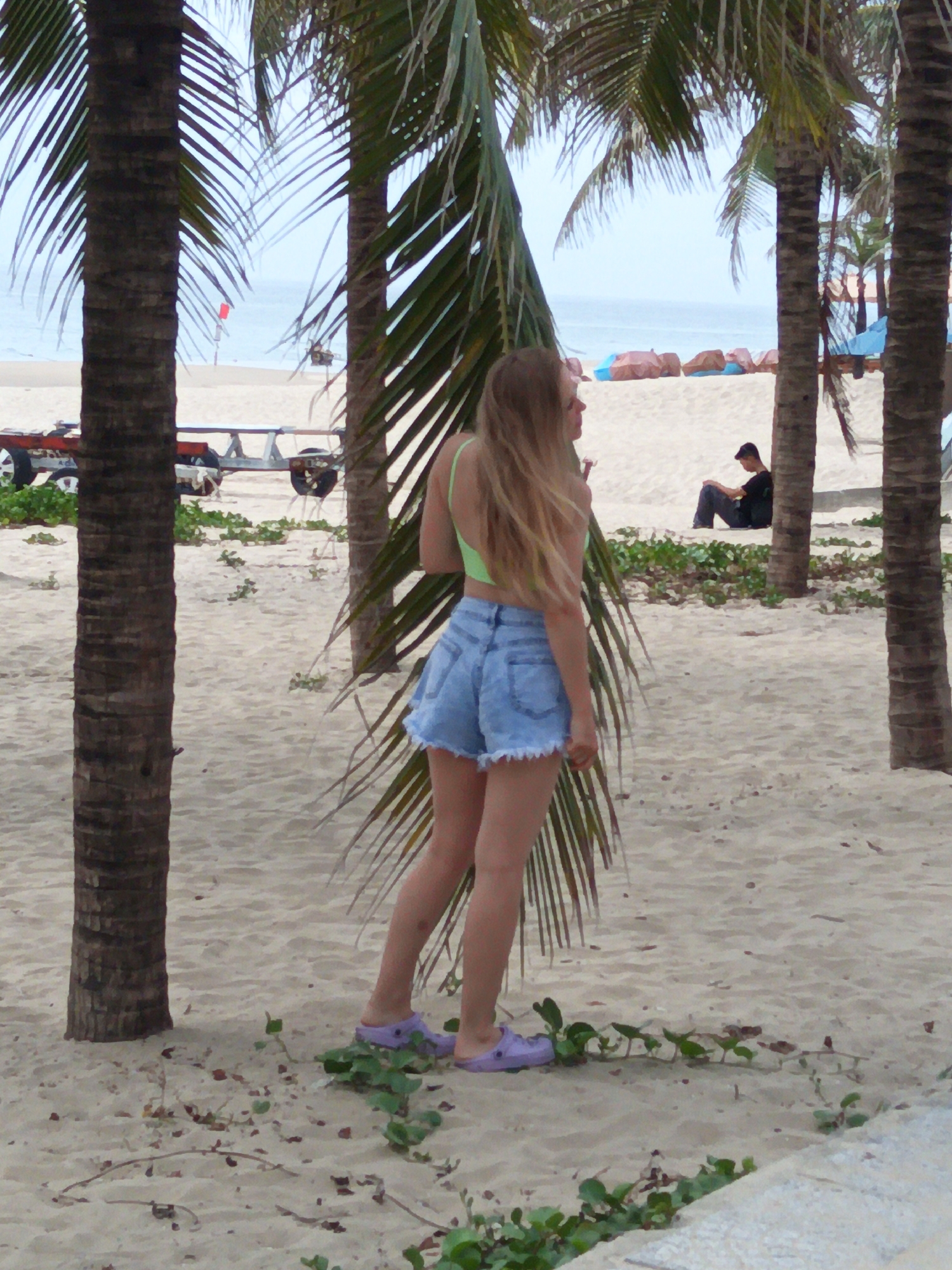 The image depicts a beach scene in Da Nang, Vietnam, with a young woman in the foreground. She is wearing a neon green bikini top and denim shorts, with light purple slip-on shoes. Her blonde hair is down, and she is looking away from the camera towards the ocean.
In the background, a man is seated on the sand, facing away from the camera. A trailer with wheels is visible on the left side of the image. Palm trees frame the scene, with their fronds partially obscuring the view. The beach is sandy, with sparse green vegetation growing in patches. The ocean and a hazy sky are visible in the distance.