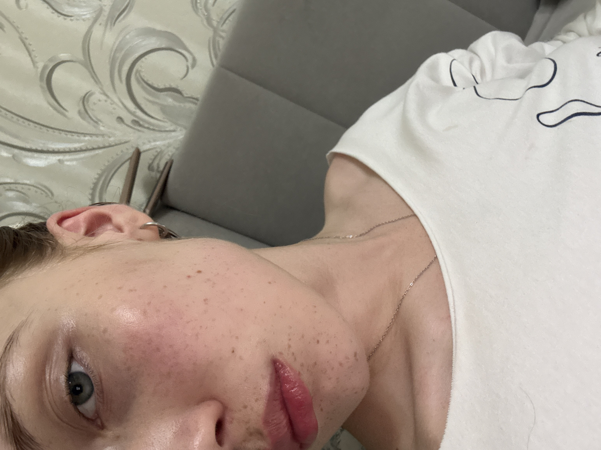 A close-up, angled portrait captures a person from the nose to the upper chest, within a domestic setting in the city of Naberezhnye Chelny, Russia. The subject, exhibiting fair skin, presents numerous scattered freckles across the visible cheek, nose, and chin areas, with a noticeable flush of redness on the left cheek. The left eye, with a blue-grey iris, is visible, showing a slight shadow beneath it. The lips are full, glossy, and tinted reddish-pink. The right ear is partially visible, adorned with a plain silver hoop earring, with dark hair pulled back from the face. The subject wears a white long-sleeved top, partially revealing a dark, abstract, curvilinear design on the fabric. A thin, silver-colored chain necklace rests on the visible skin of the neck and upper chest. The background features light-colored wallpaper with a raised, ornate, swirling floral or rococo pattern, situated above a padded headboard or sofa back upholstered in a solid grey fabric with vertical seams. The scene is illuminated by soft, ambient light.