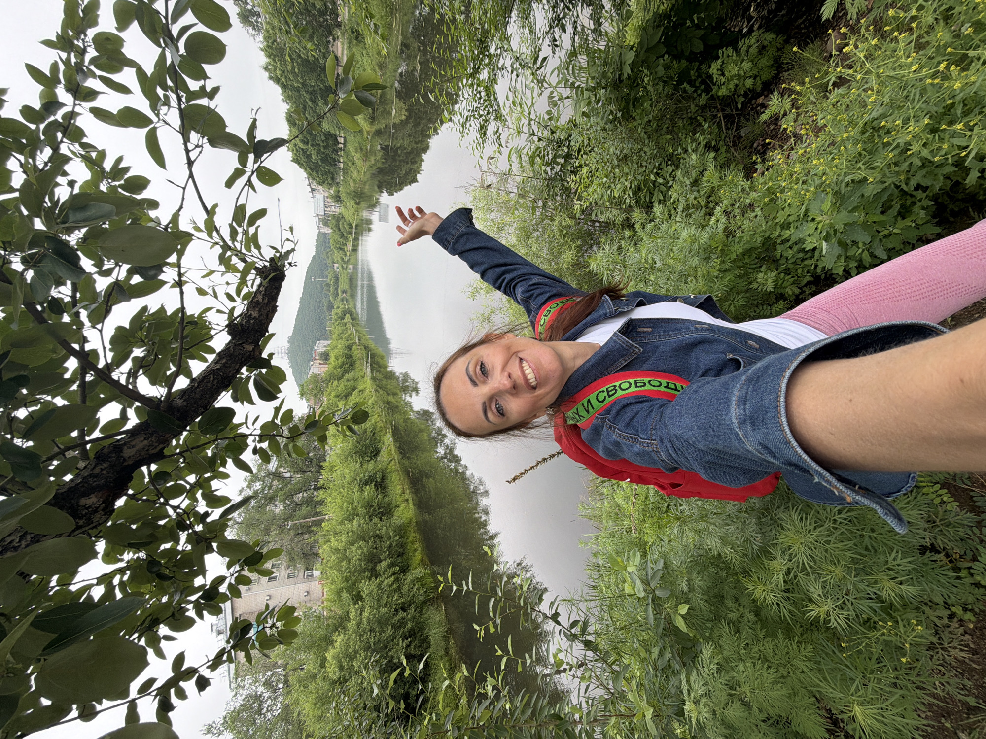 A woman with brown hair pulled back and wearing a denim jacket over a white t-shirt and pink leggings takes a selfie by a river. She has a red backpack with a green strap that has Cyrillic text visible. Her right arm is extended, and she is smiling at the camera. The river flows through a vegetated area with trees and bushes. In the background, there are buildings and hills, consistent with the urban and natural landscape of Birobidzhan, Russia. The sky is overcast.