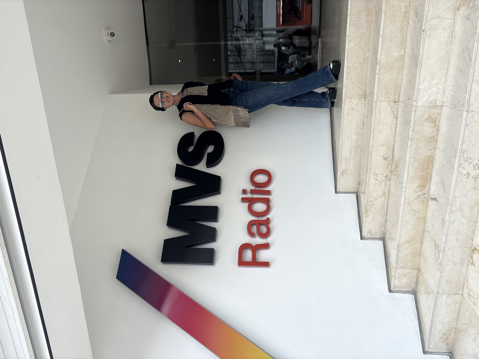 A woman stands posing in front of the entrance to MVS Radio in Mexico City, Mexico. The MVS Radio logo is displayed prominently on the wall to her left, and a marble staircase is visible on the right. The woman is wearing jeans, a black top, a beige bag, and sunglasses. She stands sideways and is facing the camera. The scene appears to be an outdoor shot taken in the daytime.