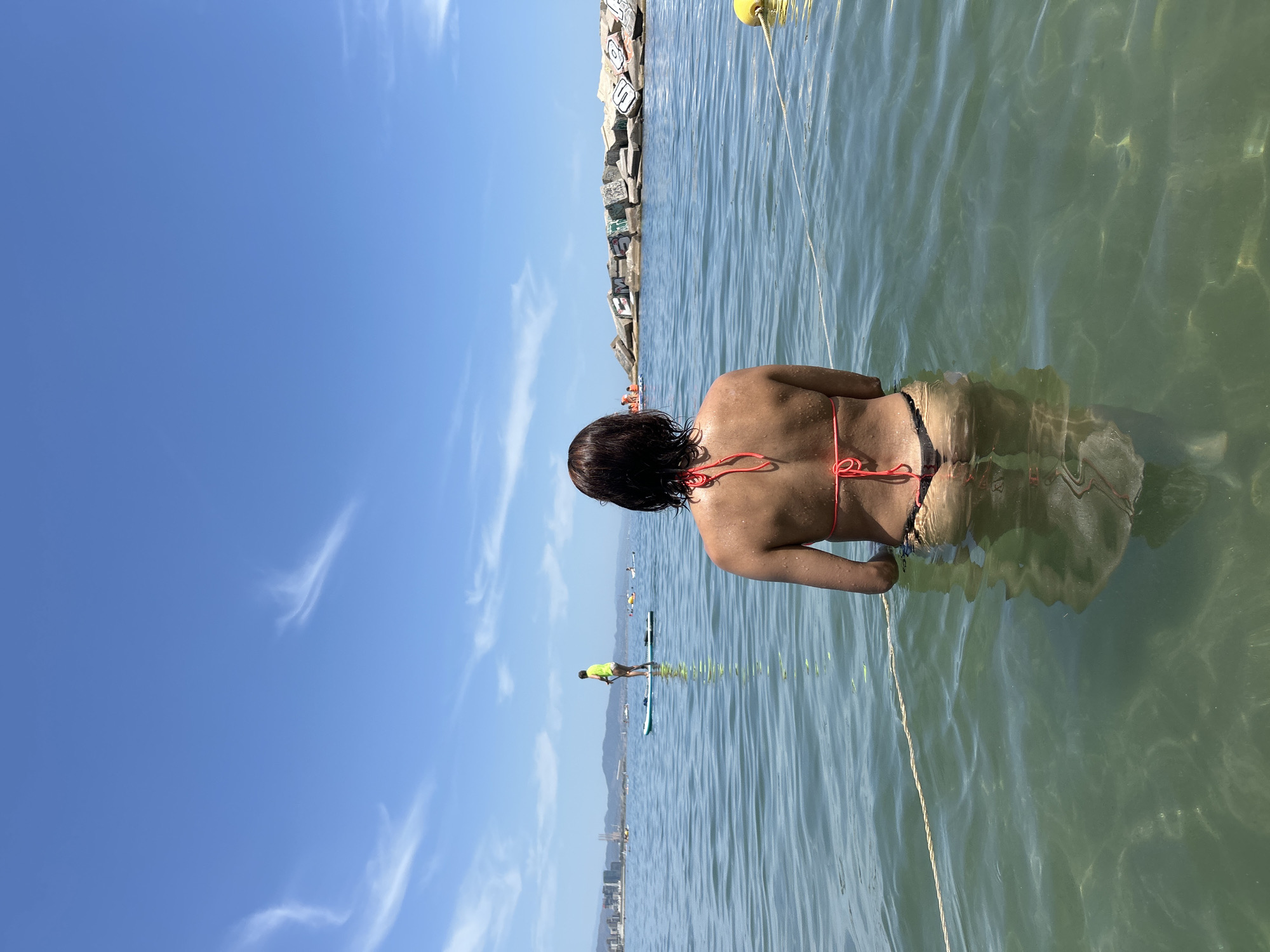 The image captures a scene at the beach in Barcelona, Spain, with a woman in a red bikini waist-deep in the ocean. She is facing away from the camera, looking out towards the water. In the distance, a person on a paddleboard can be seen, as well as a breakwater made of concrete blocks. The sky is blue with some wispy clouds. The water is clear and calm, with sunlight reflecting off the surface. The overall atmosphere is serene and summery.