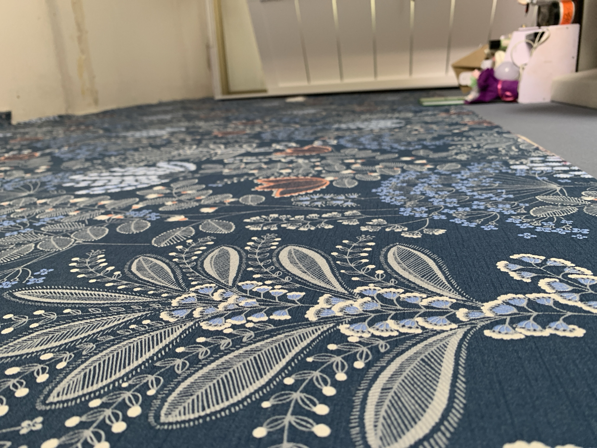 A close-up shot of a floral patterned blue floor covering, possibly a carpet or rug. The pattern consists of grey and white paisley-like motifs and blue floral designs on a dark blue background. In the background, slightly blurred, are parts of furniture or equipment, including what appears to be a white cabinet or drawer unit. The scene seems to be indoors. The image does not contain enough information to determine the location as Stavropol, Russia.