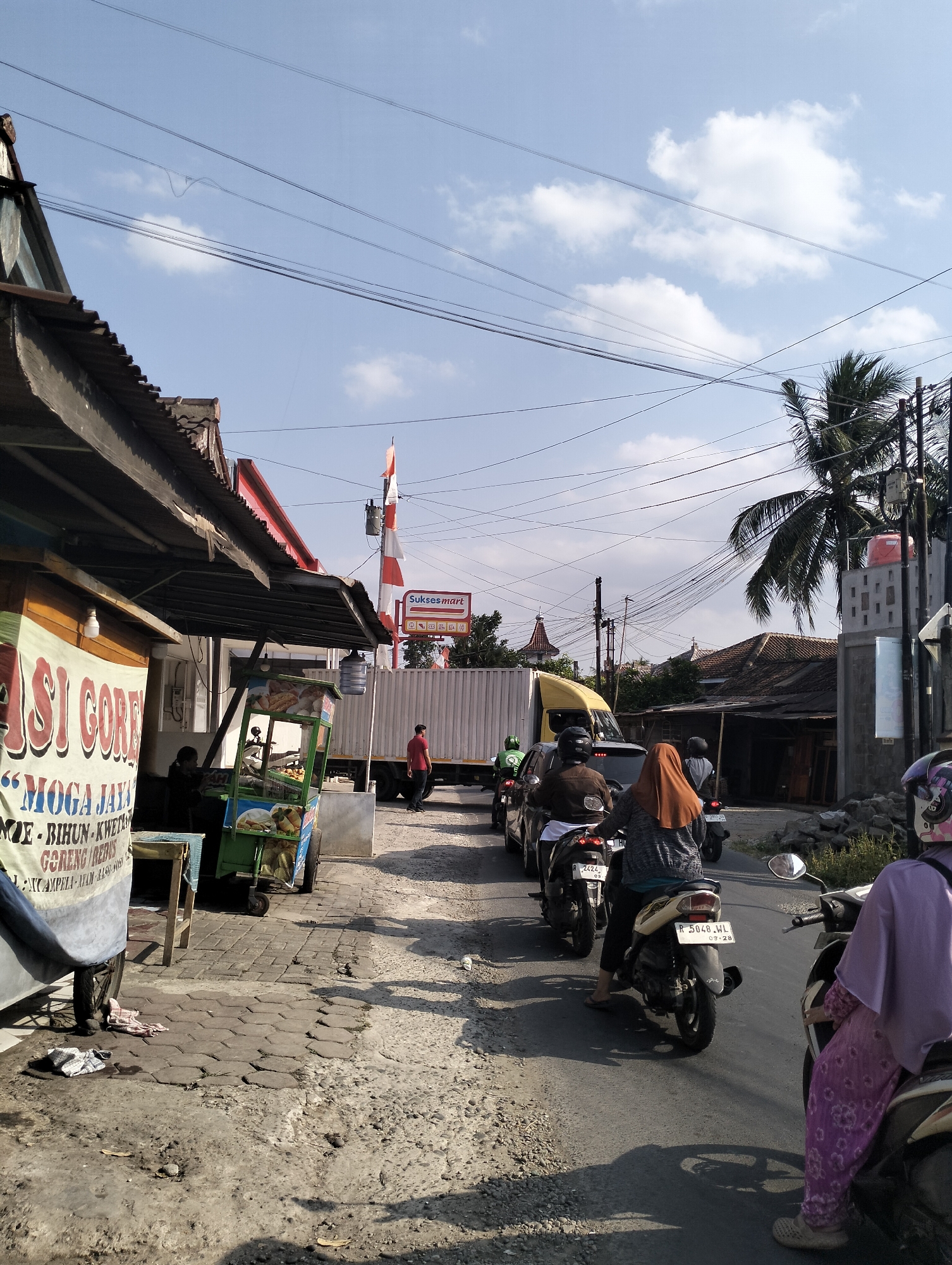 A street scene in Purbalingga, Indonesia, shows a moderately busy road with a queue of motorcycles. On the left, a food stall with a banner reading "JASI GORENG" is visible, next to a small cart selling food items. Further down the road, a large white truck is parked, and a man in a red shirt stands nearby. A red and white flag with the "Suksesmart" logo is displayed on a pole. The sky is clear blue with scattered white clouds, and a palm tree is visible on the right. The road surface is asphalt with gravel on the sides. Several people are riding motorcycles, some wearing helmets and headscarves. The overall impression is a typical day on a street in a town in Indonesia, with commercial activity and traffic.
