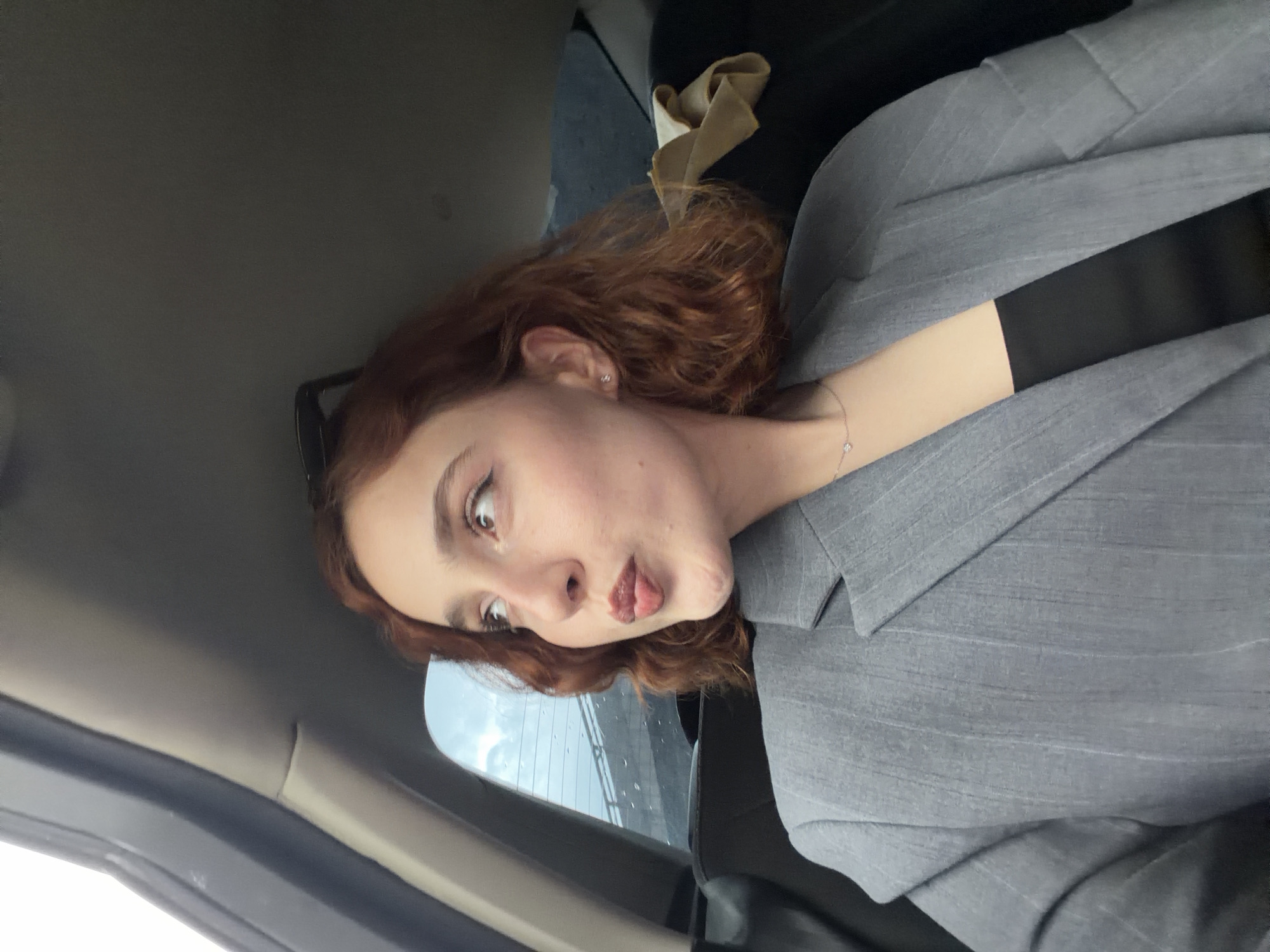 A person with auburn, curly hair and fair skin, wearing a grey pinstripe blazer and a black seatbelt, is sitting in a car. The person is making a funny face, sticking out their tongue and looking to the left. The car interior includes a light grey door and ceiling. Rain is visible outside the window. The location is likely Tbilisi, Georgia, based on the user query.