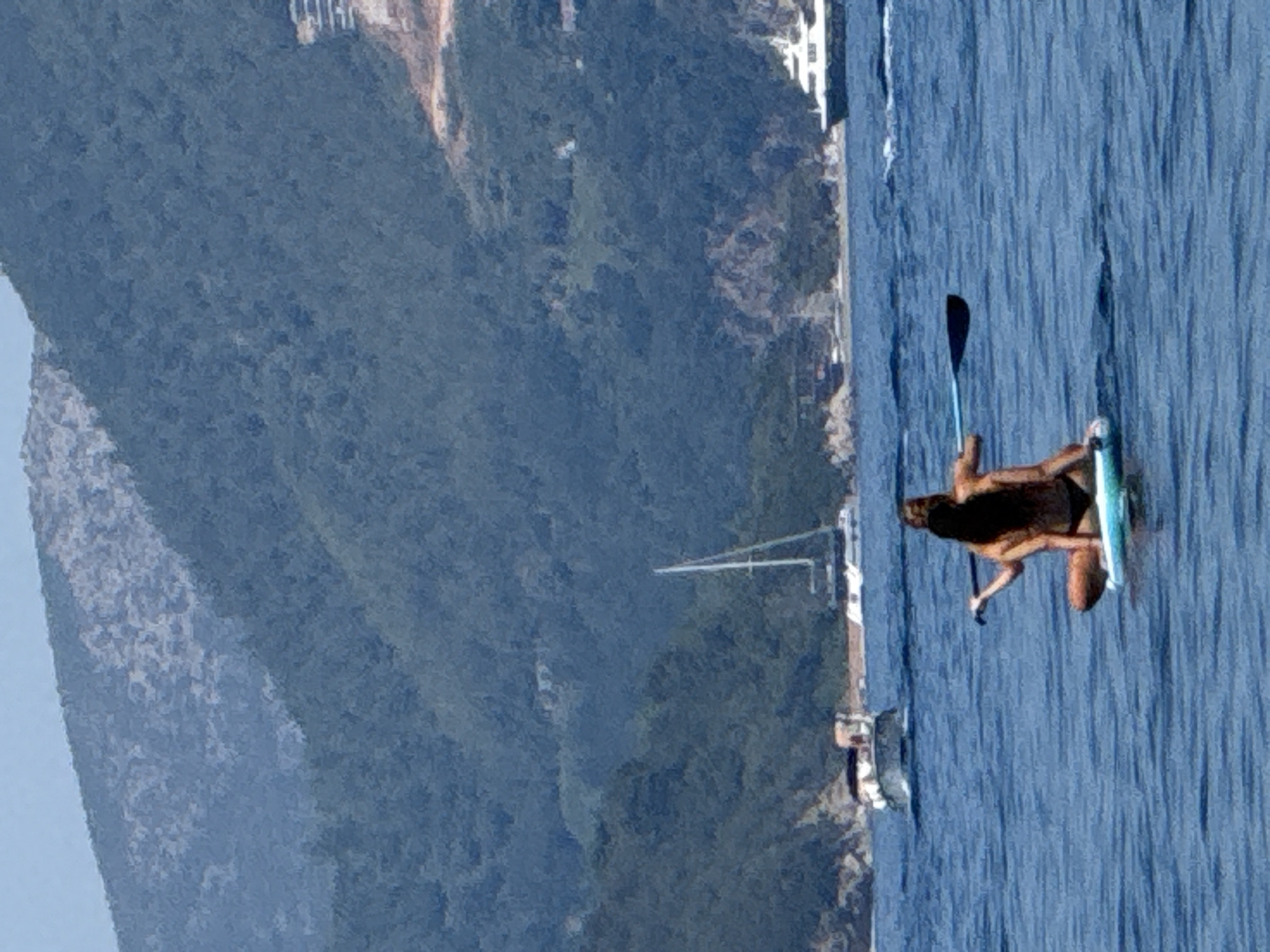 Two individuals are on a paddleboard in the water. One person is sitting cross-legged, facing away from the camera, while the other person is kneeling behind them, also facing away. The kneeling person is holding a paddle. The paddleboard is light blue. In the background, there are several boats on the water, including a sailboat and a larger white yacht. Lush green mountains rise behind the water, with some buildings visible on the slopes. The water is blue and slightly choppy. The scene appears to be a coastal area.