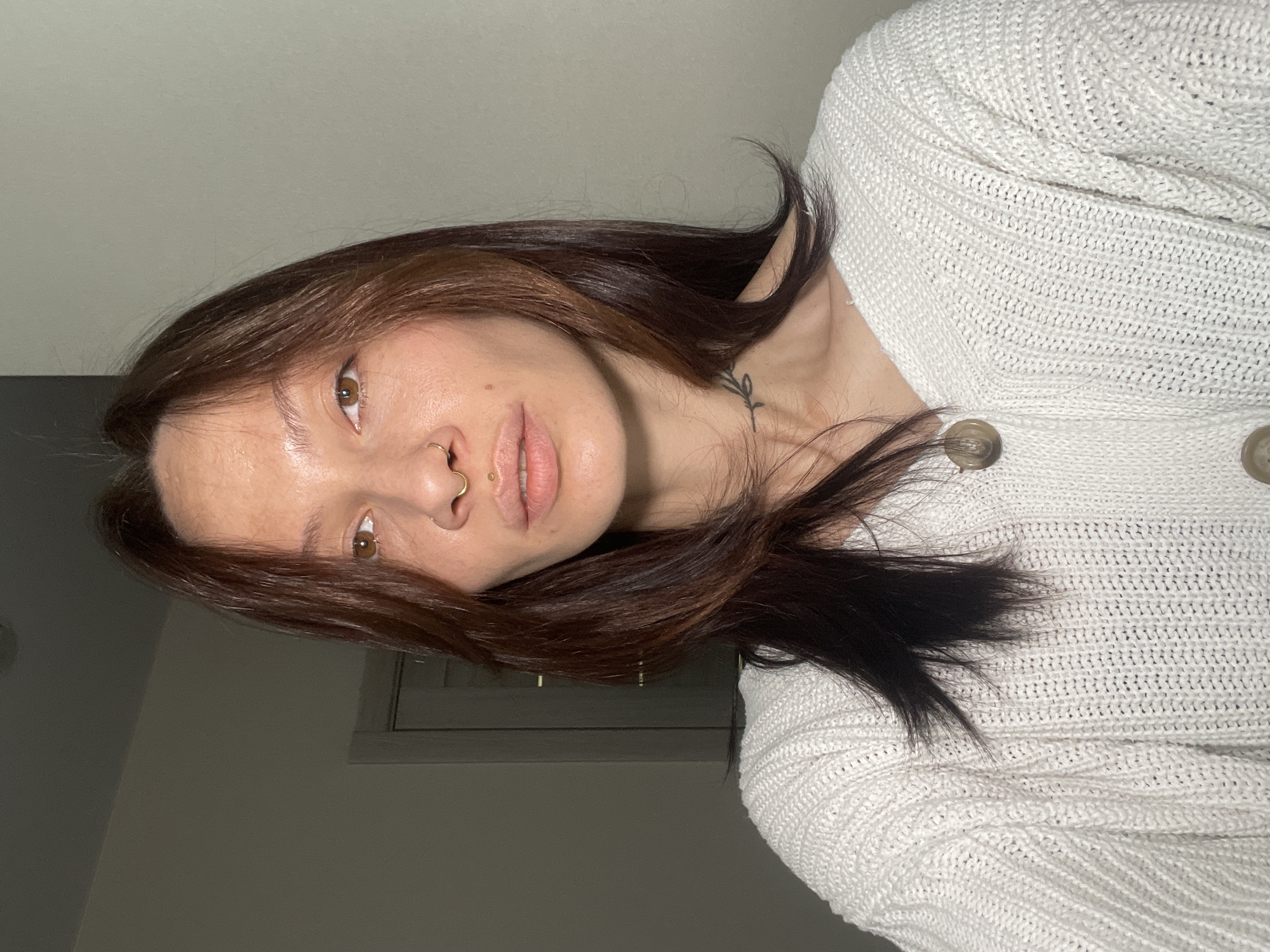 A close-up portrait features a young person, likely female, with medium-brown hair and brown eyes, looking directly at the camera. The image is oriented with the subject's head significantly tilted, giving the appearance of an intimate, angled shot.
The individual is wearing a white, chunky knit cardigan, with two light brown buttons visible on the right side of the frame. They have several piercings, including a gold-colored hoop nose ring in their right nostril and a small gold stud above their upper lip (philtrum piercing). A small, black line-art tattoo depicting a plant stem or branch with leaves is visible on the left side of their neck, near the collarbone. Their lips are slightly parted, and they maintain a neutral expression.
The background is a plain, light grey wall, indicating an indoor setting, consistent with a domestic environment in Izhevsk, Russia. A dark, rectangular shape, possibly a window frame or part of a piece of furniture, is faintly visible in the lower left portion of the image. The lighting is soft and even, illuminating the subject's features and the intricate texture of the knitwear. There is no visible text within the image.