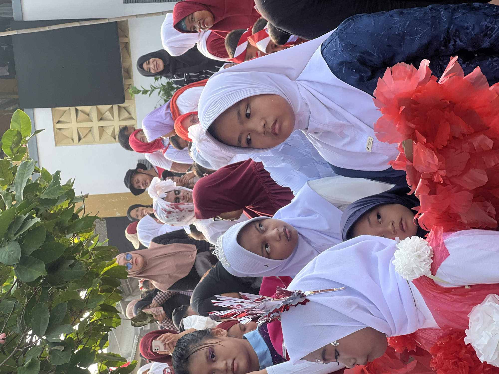 The image shows a crowd of people, primarily women and girls, in what appears to be an outdoor event. The majority are wearing headscarves, with many in hijabs. Several young girls in the foreground are wearing white hijabs and festive attire, some adorned with crowns or decorative headpieces. The girl in the center right is wearing a dark blue, embroidered outfit and a white hijab, holding a cluster of red, tissue-paper-like flowers. The girl to her left, in the foreground, is wearing a white hijab and a pink and white outfit, also decorated with red and white paper flowers and a metallic red and silver headpiece. Another girl in a white hijab with a small tiara is visible in the mid-ground, looking to her right. In the background, a person is seen wearing a white head wrap with what appears to be fake blood splattered on it, adding a potentially theatrical or themed element to the gathering. Other individuals in the background are dressed in a mix of casual wear and more traditional clothing. The overall impression is of a public gathering, possibly a parade, festival, or cultural event. There are elements of green foliage from a tree in the upper left corner, suggesting an outdoor setting. The background includes architectural elements, with a white wall featuring a geometric pattern and a large dark rectangular object. The presence of red and white decorations, particularly the flowers and headbands on some individuals, could indicate a patriotic theme, possibly related to Indonesian Independence Day. The scene is well-lit, suggesting daytime. The context of Klapanunggal, Indonesia, implies this is likely a local community event.