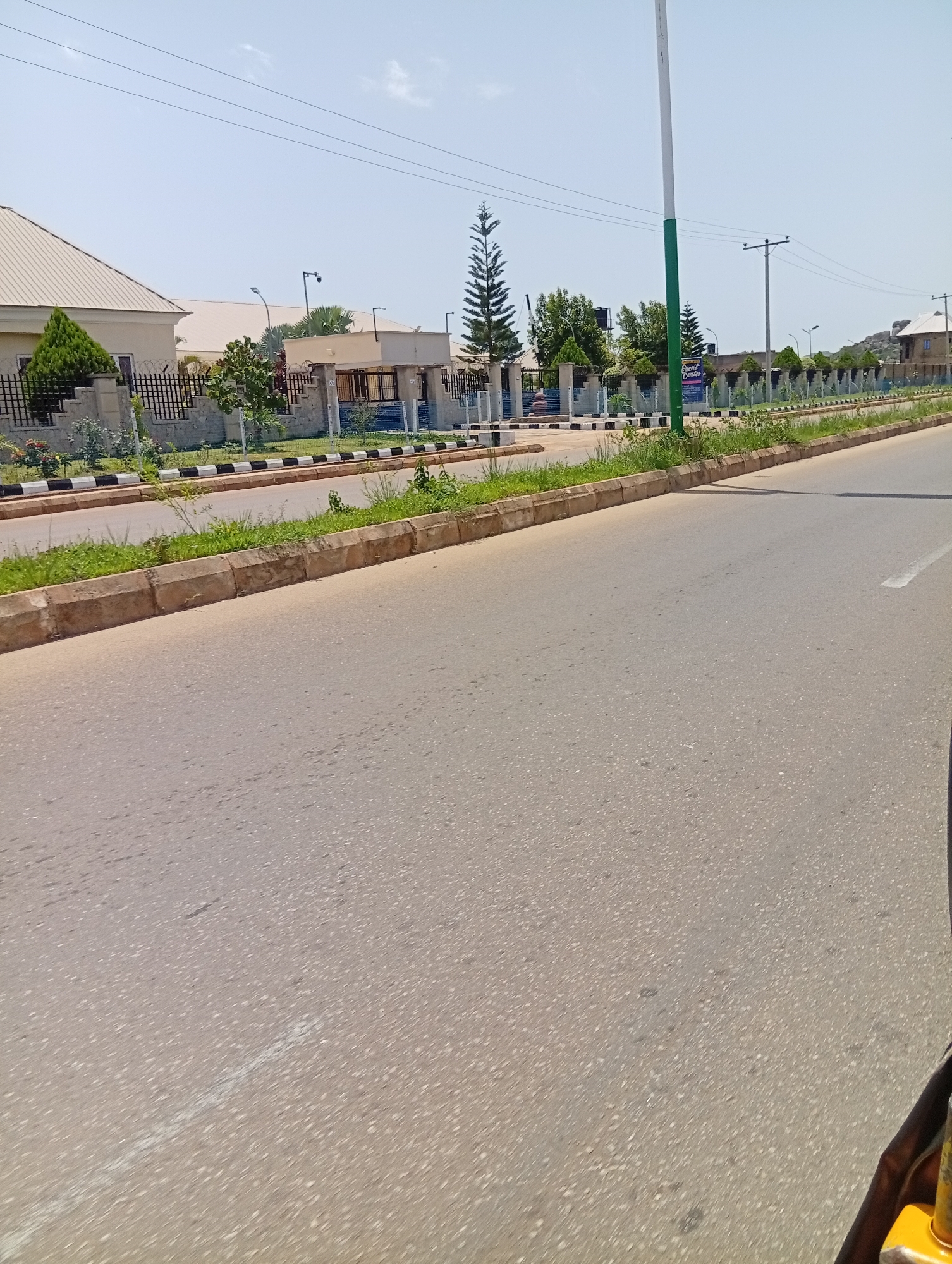 The image depicts a roadside scene in Jos, Nigeria, characterized by an asphalt road in the foreground, a concrete curb, and a strip of green grass and vegetation bordering the road. Beyond the vegetation, a black metal fence encloses a property with a beige, gabled-roof building visible on the left. Further along the road, a tall, slender evergreen tree stands out, accompanied by other trees and smaller shrubs. Several utility poles with power lines are also visible in the background, along with a tall white and green pole. The sky is clear and bright. The scene appears to be a residential or commercial area with well-maintained landscaping.