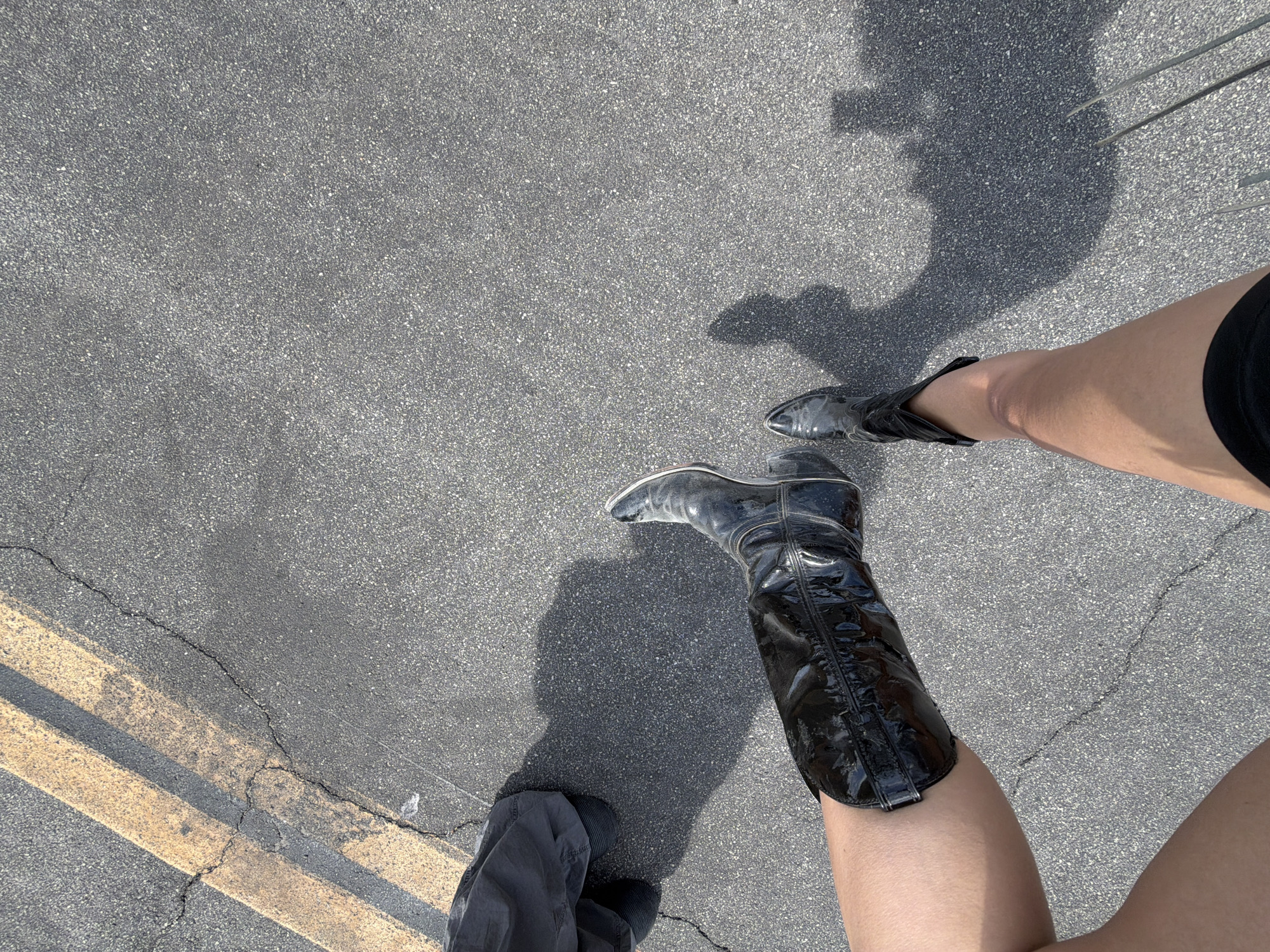 The image shows two people standing on asphalt. One person is wearing a shiny, black, knee-high boot on their right leg and gray pants. The other person is wearing a black shoe. The location is likely a street or parking lot in Palm Desert, California, United States of America. Evidence of this assumption includes asphalt ground that is typical of many streets and parking lots, and the asphalt having yellow pavement lines. There is a shadow that indicates the time of day is during daylight hours.