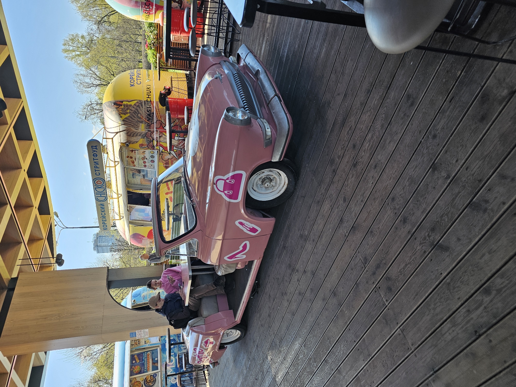 The image shows a pink vintage car that has been repurposed as a dining area. The car is decorated with stickers, including a handbag logo and the number "2". People are seated inside the car, using it as a place to eat or relax. In the background, there is a yellow food truck advertising "Korean street food". The scene appears to be set in an urban outdoor area, possibly a street or a patio, as suggested by the wooden flooring and the presence of other tables and chairs. Based on the Cyrillic script on the food truck, it's most likely somewhere in Moscow, Russia.