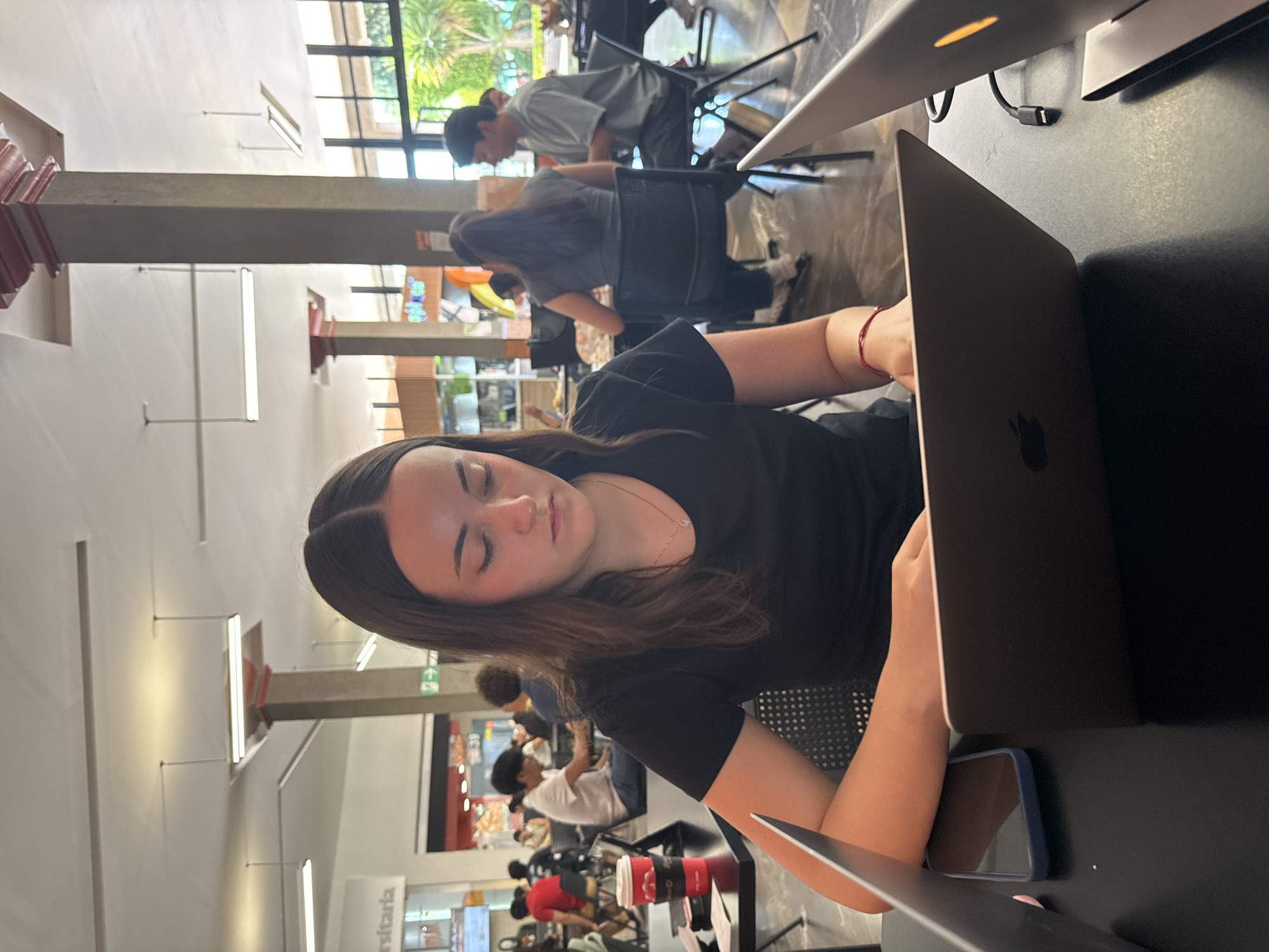 The image depicts an indoor scene, likely a university library, study hall, or cafe, in San Andrés Cholula, Mexico, during daylight hours.
The primary subject is a young woman with long, dark hair, wearing a black t-shirt, seated at a dark table. She is focused on a silver Apple laptop, with her hands poised above the keyboard. A red string bracelet is visible on her right wrist. Next to her laptop, a smartphone in a blue case stands upright on the table.
In the midground and background, several other individuals are present. Some are seated at tables, also using laptops or engaging in conversations, while others are standing. A man in a light grey t-shirt is visible near a large window. These windows feature dark frames and offer a view of lush green foliage outside, indicating a bright and clear day. The interior space features modern architectural elements, including concrete pillars and high ceilings equipped with linear fluorescent lighting fixtures.
On a wall or counter in the background, a white sign is partially visible, bearing the Spanish word "Universitaria," which strongly suggests a university environment. Red coffee cups are also seen on a distant table. The overall atmosphere is one of focused activity in a well-lit, contemporary setting.