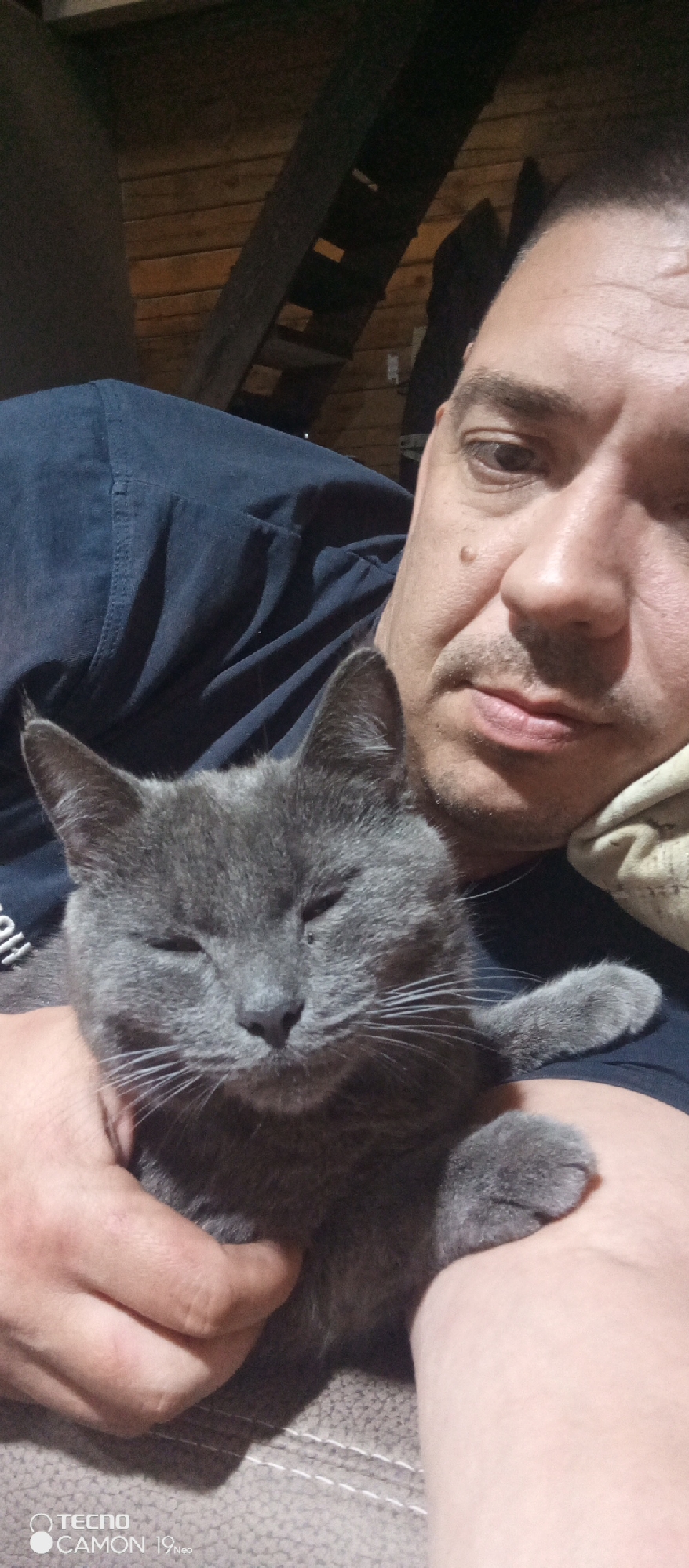 A man is holding a gray cat. The location is in a room with wooden walls, possibly in Chaikovsky, Russia.