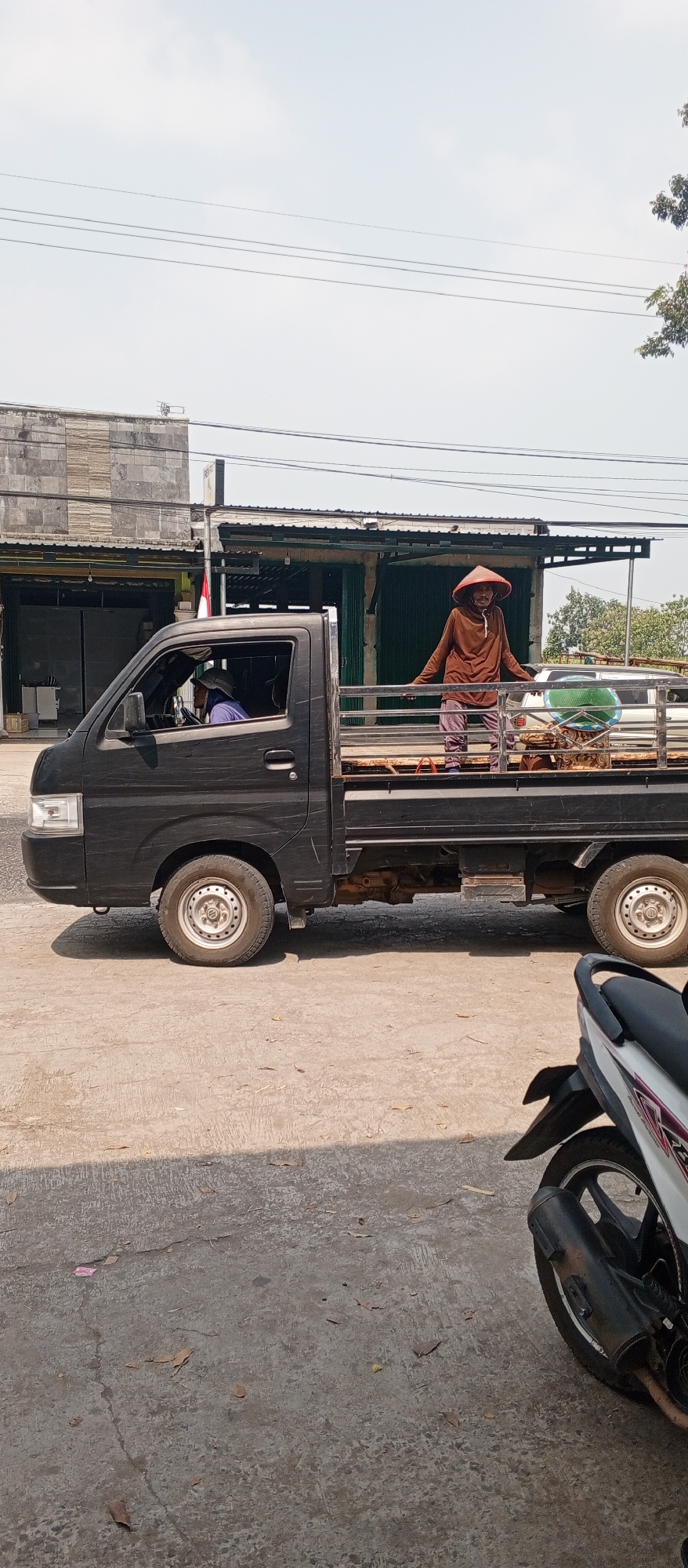 In the city of Maospati, Indonesia, a black pickup truck is parked on a paved road. The truck is carrying two people: a person wearing a conical hat and a brown long-sleeved shirt, standing in the back of the truck, and another person partially visible in the driver's seat, wearing a light-colored hat and a purple shirt. The person in the back of the truck appears to be interacting with something on the truck bed. The truck bed contains various items, including what looks like woven mats and a green basket. The environment includes buildings with metal roll-up doors and trees in the background. A motorcycle is parked in the foreground, partially obscuring the truck. The sky is visible and appears clear.