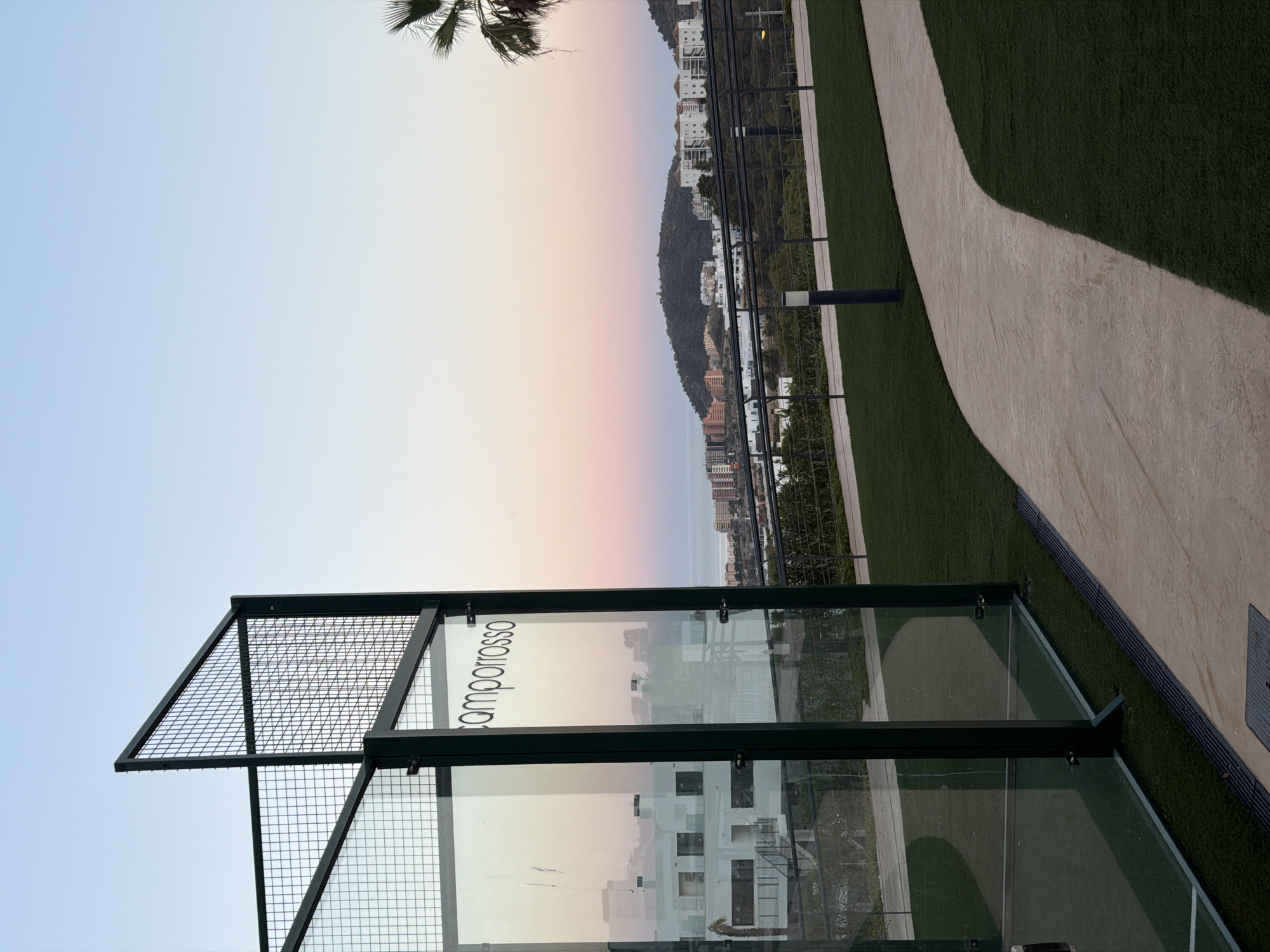 An outdoor scene at dusk features a paddle tennis court in the foreground, with its glass walls and metal netting visible on the left. A beige pathway curves through artificial green turf on the right, leading towards a distant cityscape and hills. The sky displays a gradient of pale blue to pink, indicative of sunset. Buildings are scattered across the landscape, with a prominent hill topped by structures in the mid-ground. A palm tree frond is visible on the far right. The location is identified as the city of Finestrat, Spain. There are no people visible in the image.
