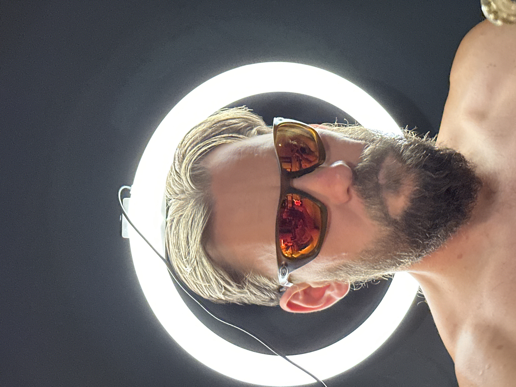 A man with blond hair and a beard is wearing sunglasses and standing in front of a ring light. The location is not discernible from the image.