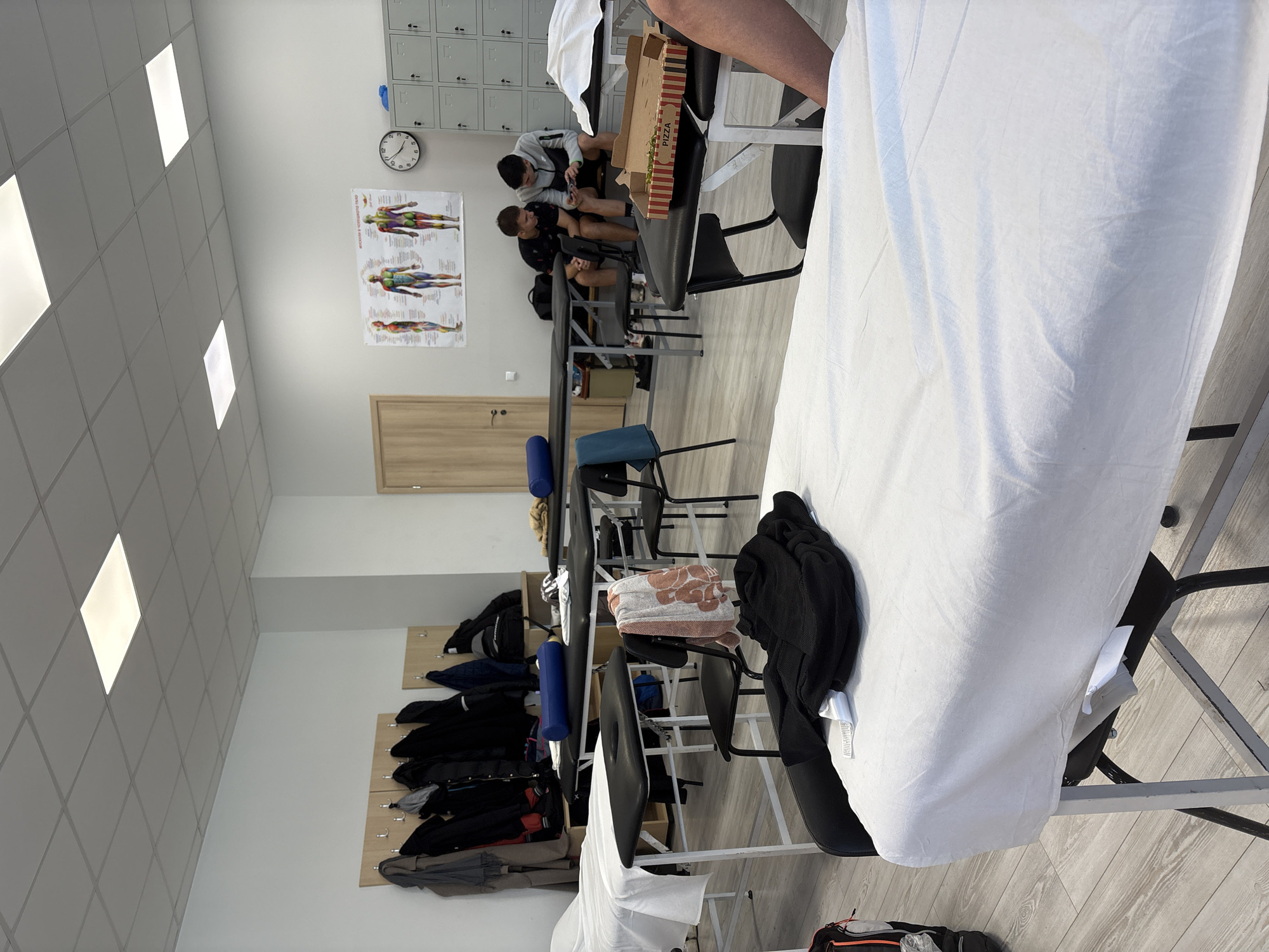 The image depicts a room that seems to be part of a sports or medical facility. Two young men sit in chairs, looking at a mobile phone, positioned near lockers on a wall. A pizza box sits on a chair near them, partially open. A medical poster or chart hangs on the wall above a closed light brown door. Further into the room, chairs are scattered, and a wall-mounted coat rack holds numerous coats. In the foreground, a medical bed covered with a white sheet is in focus. A black jacket and a patterned towel are draped over chairs nearby. The room is brightly lit by square ceiling lights. The location is assumed to be in Sofia, Bulgaria, based on information external to the image.