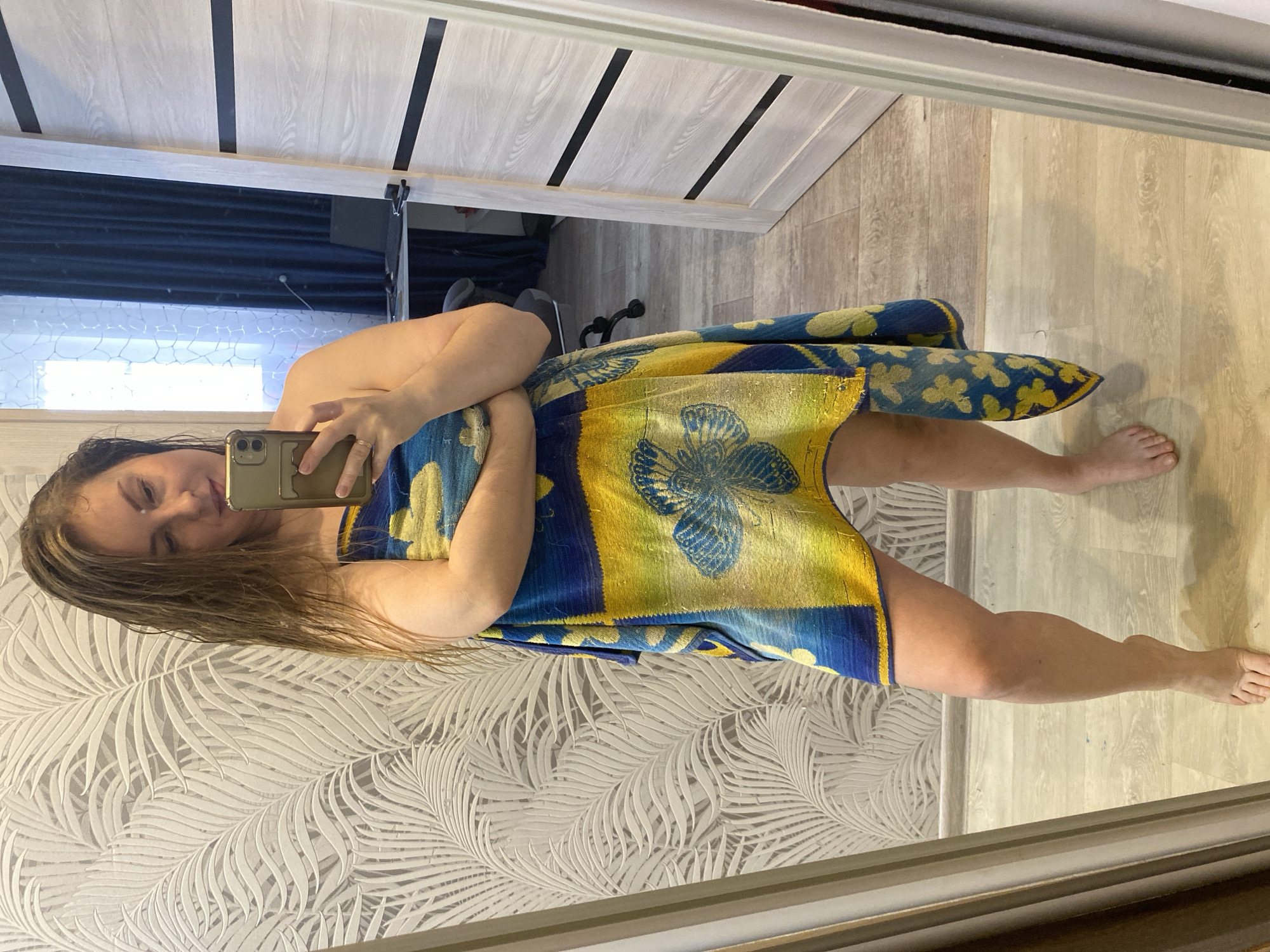 A woman takes a mirror selfie indoors. She is seen from the chest up, with a blue and yellow towel, featuring a butterfly and floral pattern, wrapped around her body, covering her from just above her chest to mid-thigh. Her hair appears damp or wet, suggesting she may have recently showered. She holds a dark-colored smartphone, possibly an iPhone, with multiple rear cameras, to her face, capturing her reflection. Her expression is neutral with a slight smile. A ring is visible on her right hand.
The reflection in the mirror reveals the surrounding room. To the left, a light-colored wallpaper with a white-on-white fern or palm leaf pattern is visible. The floor is covered with light wood-look laminate or planks. Behind the woman, a light-colored door with dark horizontal stripe accents is reflected. In the background of the reflection, a dark blue curtain and what appears to be a window with a pattern (possibly frost or a net curtain) are visible. No explicit text is visible in the image. The setting appears to be a private residence.