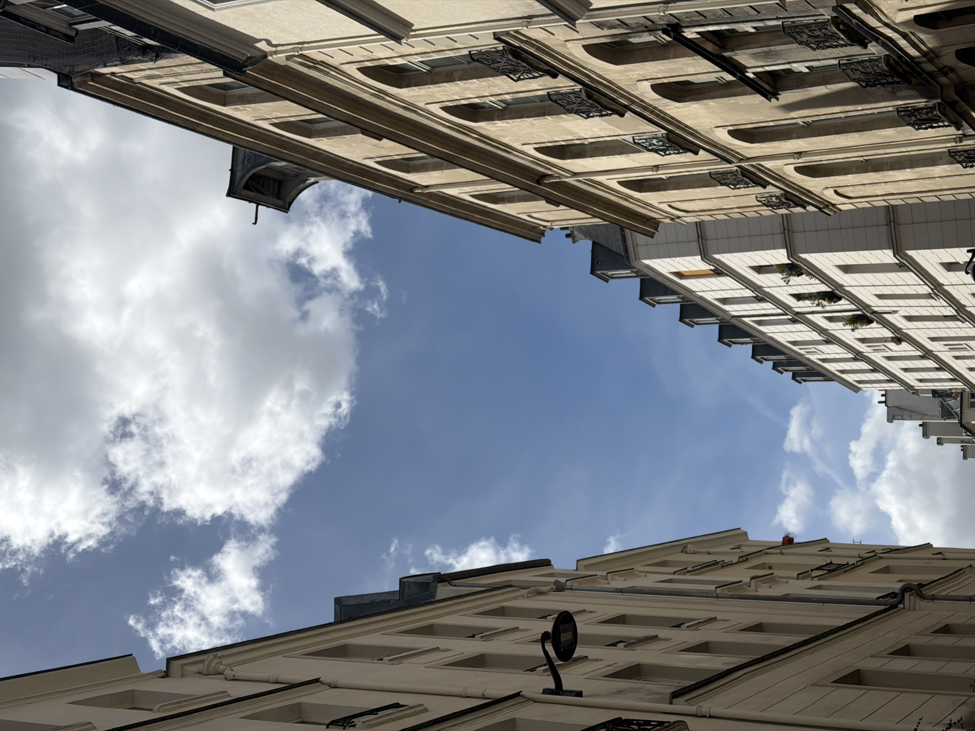 The image presents an upward perspective of tall, classic Parisian buildings framing a clear blue sky. The scene is dominated by light-colored (cream or beige) facades rising on the left, right, and bottom edges of the frame, converging to create a V-shape of sky in the center.
The buildings feature distinct architectural details characteristic of Paris, including numerous rectangular windows with decorative frames, prominent cornices, and ornate black wrought-iron balcony railings on several floors. The facades show a structured and symmetrical design, typical of Haussmannian architecture.
On the bottom right building, a black, circular object, possibly a street lamp or a camera, is mounted on the wall. A small, indistinct red object can be seen on the rooftop at the very top of one of the buildings. Further to the right, a building with a slightly different, possibly more modern, white-paneled facade is visible, featuring regularly spaced windows, some of which contain small green plants.
The sky occupying the central space is a vibrant blue, dotted with scattered, fluffy white cumulus clouds. The bright and clear weather conditions suggest it is daytime. There are no people visible, and no specific activity is taking place. No text is discernible anywhere in the image. The composition emphasizes the grandeur of the urban structures against the vastness of the sky.