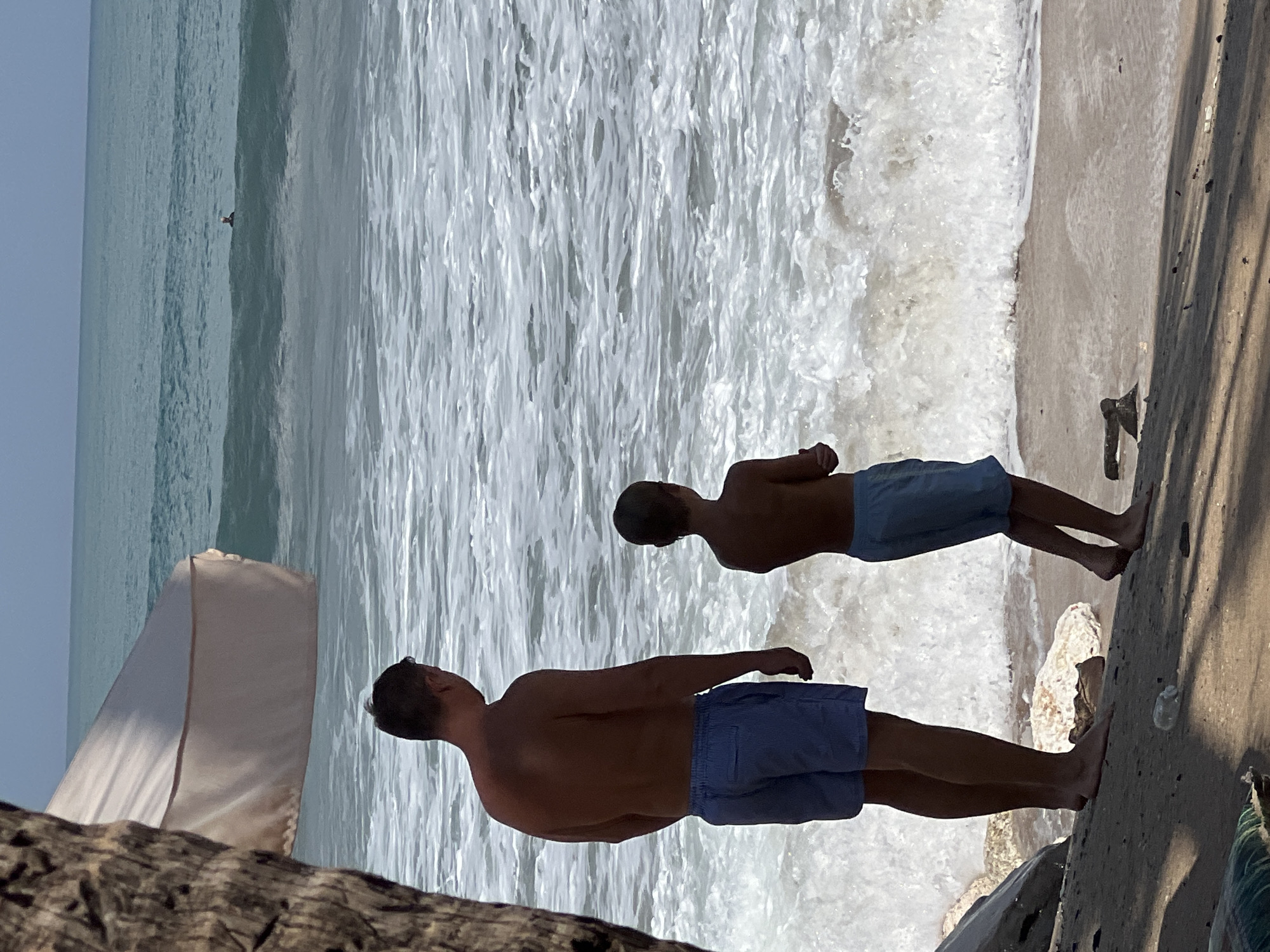 In Kuta, Indonesia, a man and a boy stand on a sandy beach, facing the ocean. The man, wearing blue striped swim trunks, stands to the left. To his right, a boy, also in blue swim trunks, stands slightly ahead. The ocean is a deep blue with white-capped waves rolling towards the shore. In the distance, a single person is visible in the water, appearing to be swimming or surfing. A large, textured object, possibly a palm tree trunk, is partially visible on the left side of the frame, and a light-colored canopy extends from the top left corner. The sand is a light brown, with scattered rocks and a plastic bottle visible. The sunlight casts shadows on the beach, indicating it is daytime.