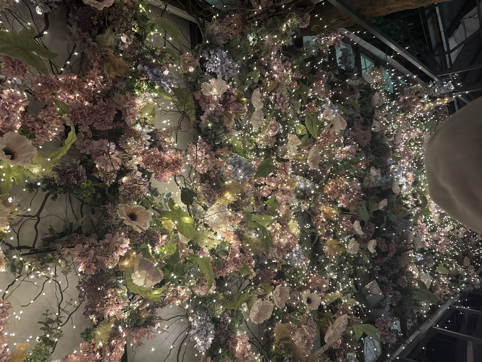 The image shows a wall covered in artificial flowers and string lights. The flowers are a mix of pinks, blues, purples, and whites, with green foliage interspersed. Thin, bare branches are also visible. The lights are small and create a twinkling effect. In the upper right corner of the frame is a dark metal structure, possibly a trellis or part of a building frame. A blurred shape, possibly a person's head, is in the far right of the frame. The image provides no information about Sofia, Bulgaria.
