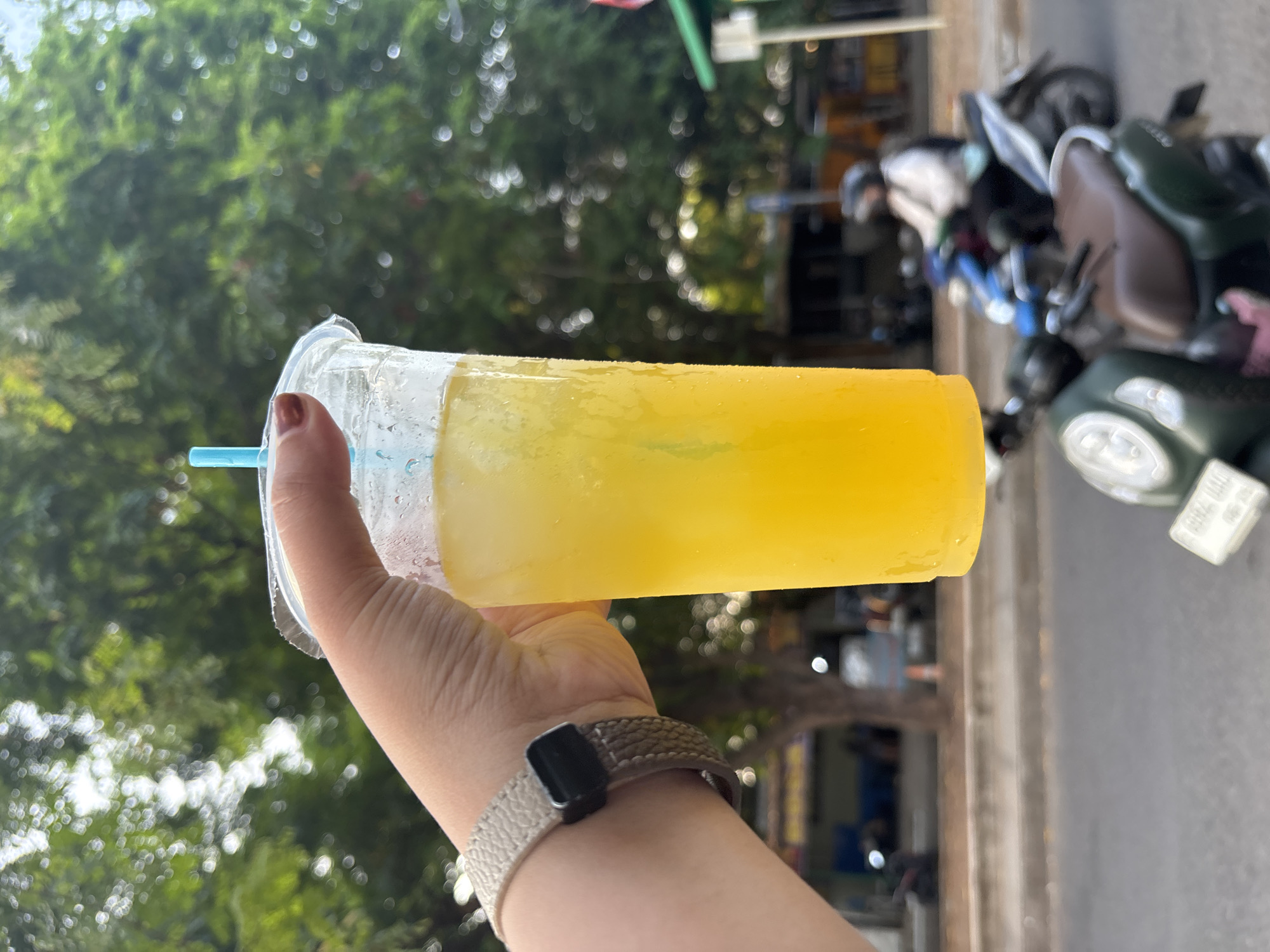 A person's hand, adorned with a light brown textured watch, holds a tall, clear plastic cup filled with a bright yellow, ice-filled beverage and a blue straw. The cup is partially obscured by the hand. The background is blurred but suggests an outdoor urban setting with lush green trees and dappled sunlight. In the middle ground, a street is visible with a sidewalk and some parked motorcycles, one of which has a rider wearing a light-colored jacket and a helmet. The scene appears to be in Cirebon, Indonesia, given the context provided.