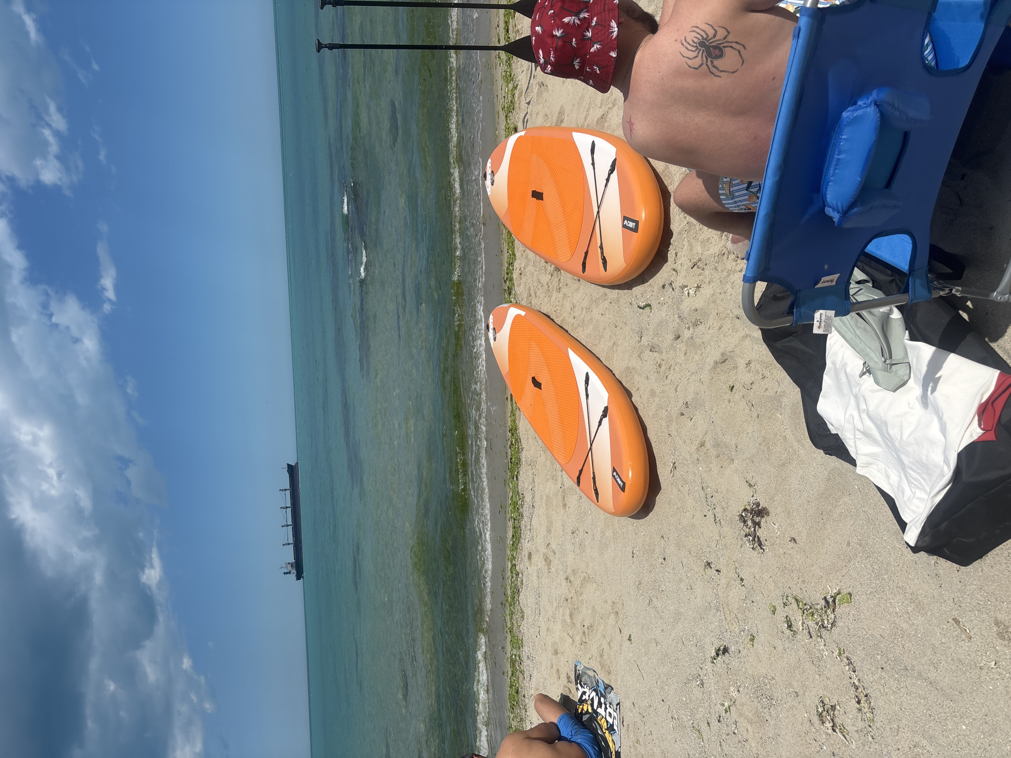 The image depicts a sandy beach scene with the sea in the background. Two orange paddleboards are lying on the sand, positioned side-by-side. In the foreground, a person is seated in a blue beach chair, facing away from the camera, wearing a patterned bucket hat. Another person is seated on the sand to the left, also facing away from the camera, wearing blue shorts. The sea is calm with a few waves breaking near the shore, and a large cargo ship is visible in the distance on the horizon. The sky is blue with scattered clouds. This scene is consistent with the beaches found in Varna, Bulgaria, known for its coastal resorts and recreational activities.