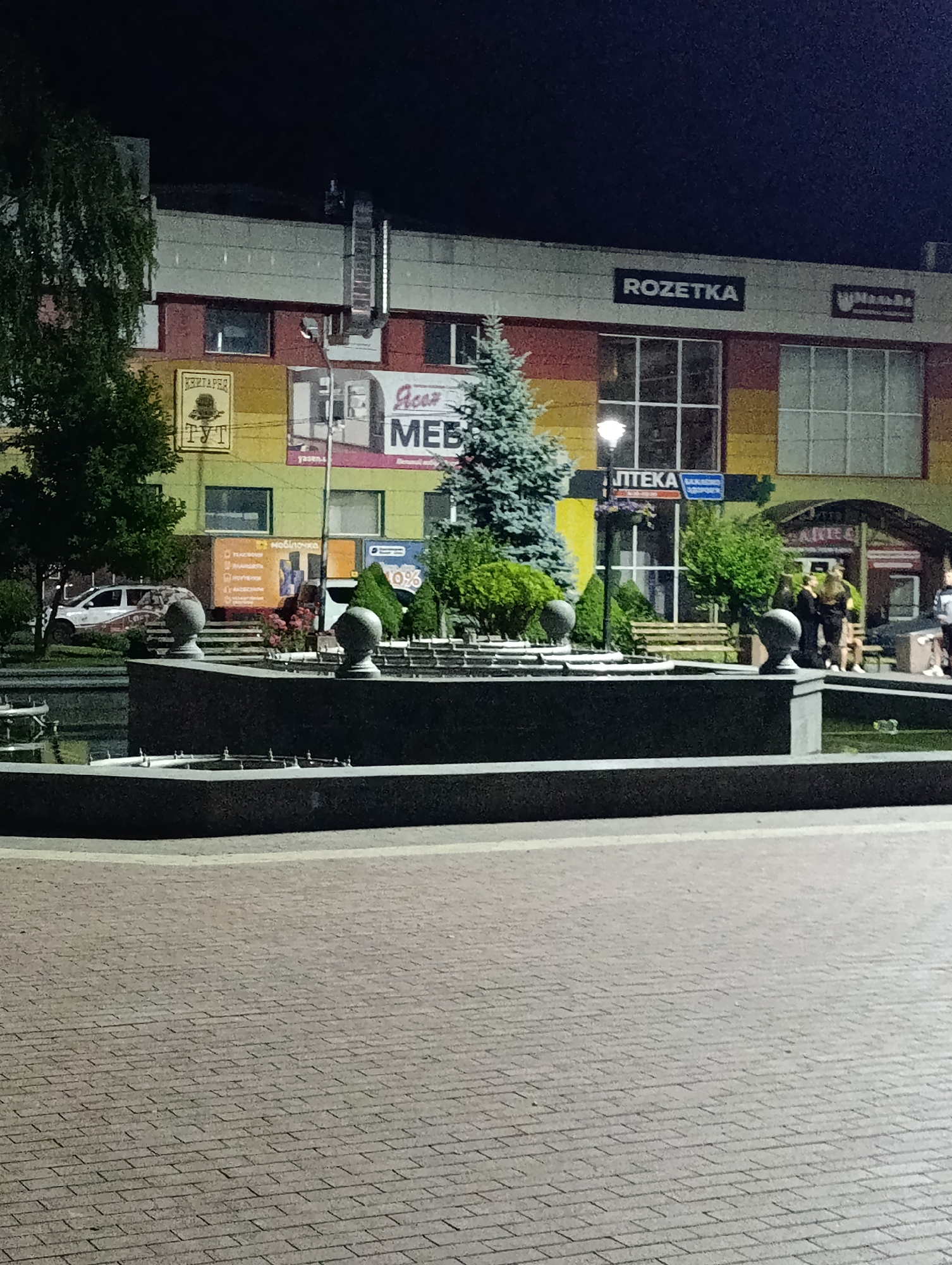 This image captures a night-time view of a commercial building in Ladyzhyn, Ukraine. The building features a multi-colored facade with sections of yellow, orange, and red. Prominently displayed on the building is the "ROZETKA" logo in black lettering on a white background. Below this, there are large windows and various advertisements, including one for "MEB" which likely refers to furniture.
In the foreground, a dark, angular fountain structure is visible, with decorative spheres on its edges. The area surrounding the fountain is paved with brickwork, with a slightly raised edge. To the left of the fountain, a tree with dark green foliage is present. Behind the fountain, a few individuals are standing and conversing near an entrance, with some seated on benches in the background. A white van is parked to the left of the building. The overall scene is illuminated by streetlights and the lights from within the commercial establishment.